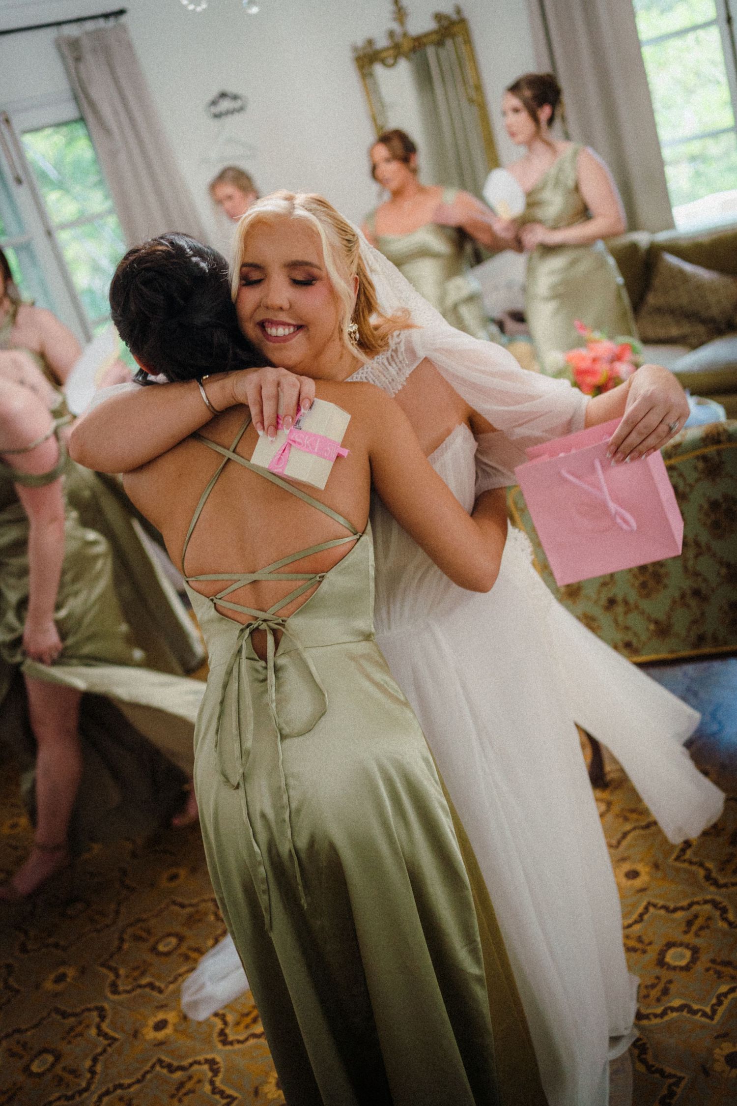 Bride in white gown hugging bridesmaid in sage green satin dress while holding pink gift bag at wedding.