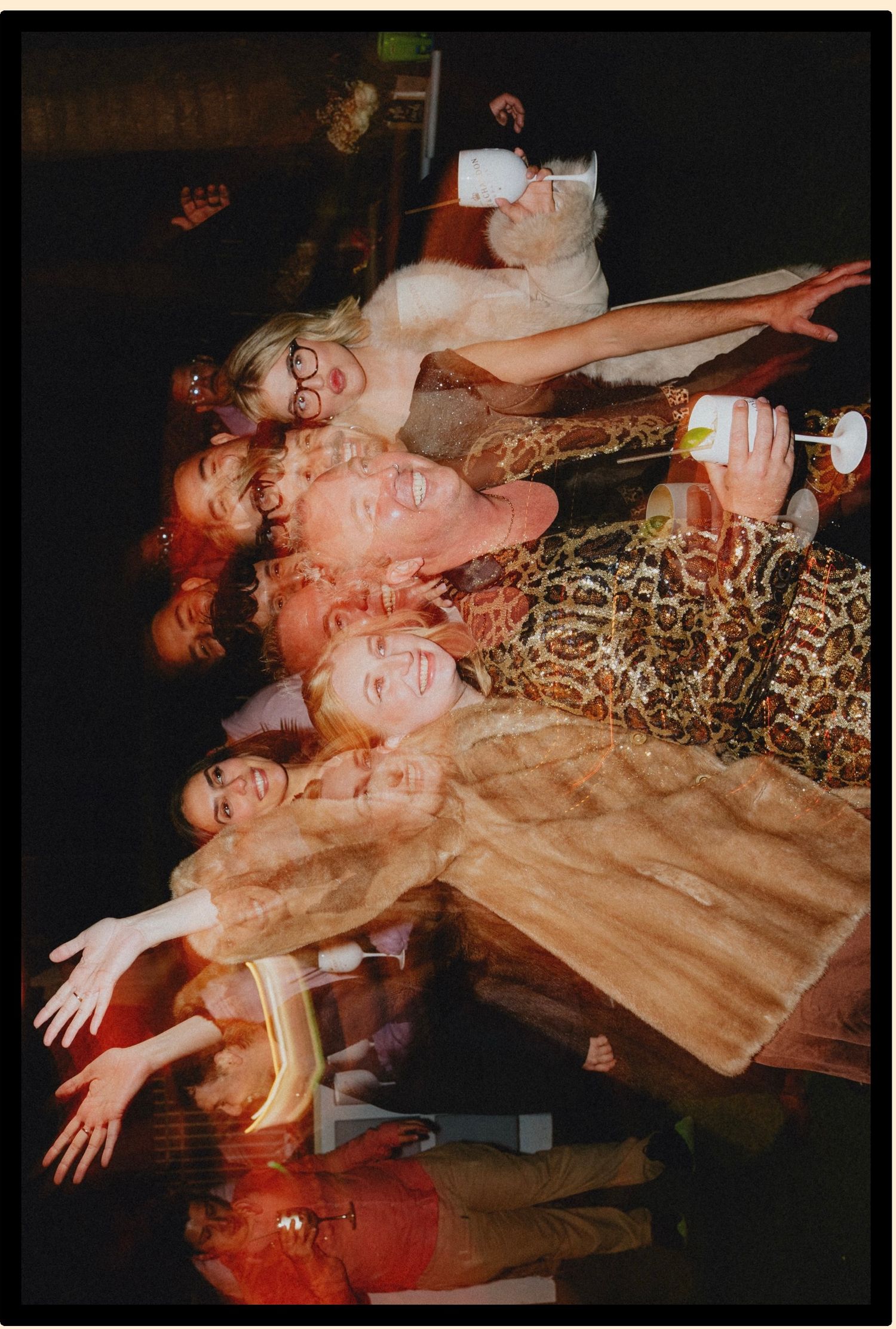 Group of friends posing together at a party, wearing colorful outfits and fur coats, holding drinks and smiling.