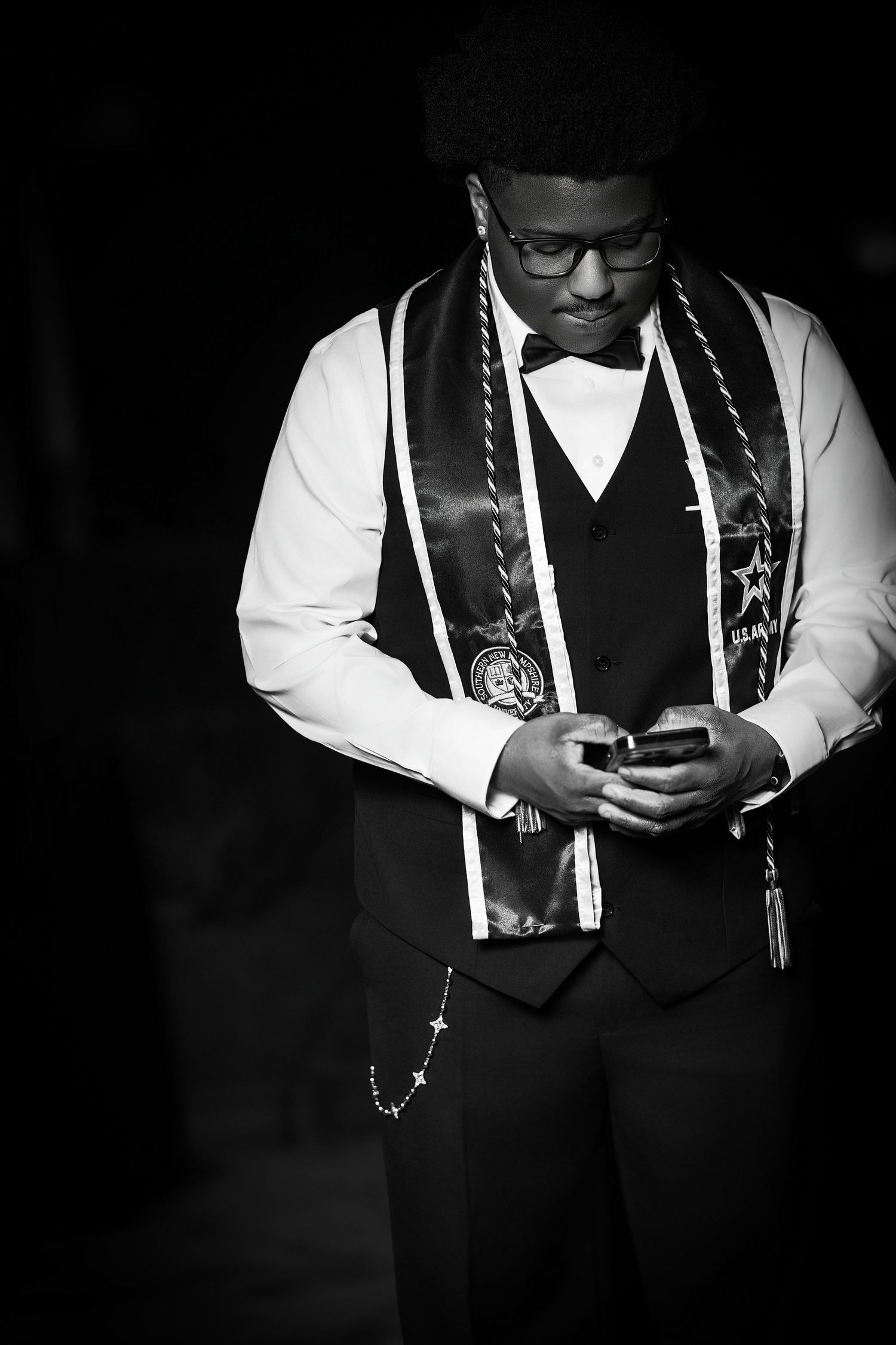 Black and white photo of a young man in formal attire with graduation sash checking his phone.