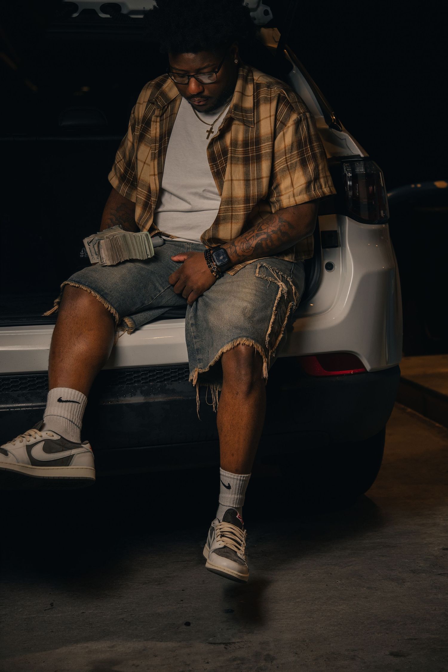 Man in plaid shirt, denim cutoffs, and Nike Air Jordan 1s sits on vehicle tailgate in dimly lit garage.