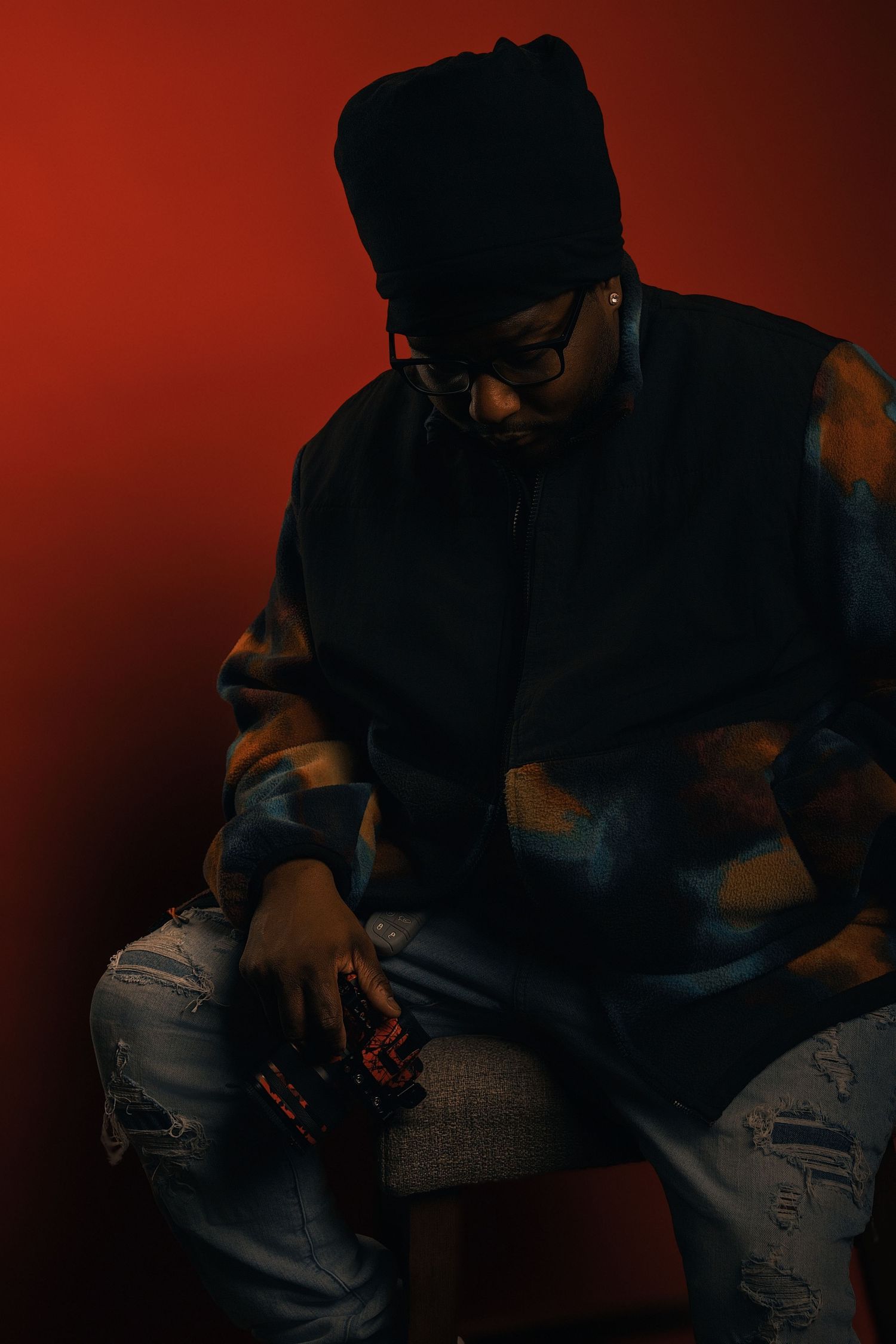 Young man in black beanie, glasses, and ripped jeans sits against a red background in dramatic lighting.