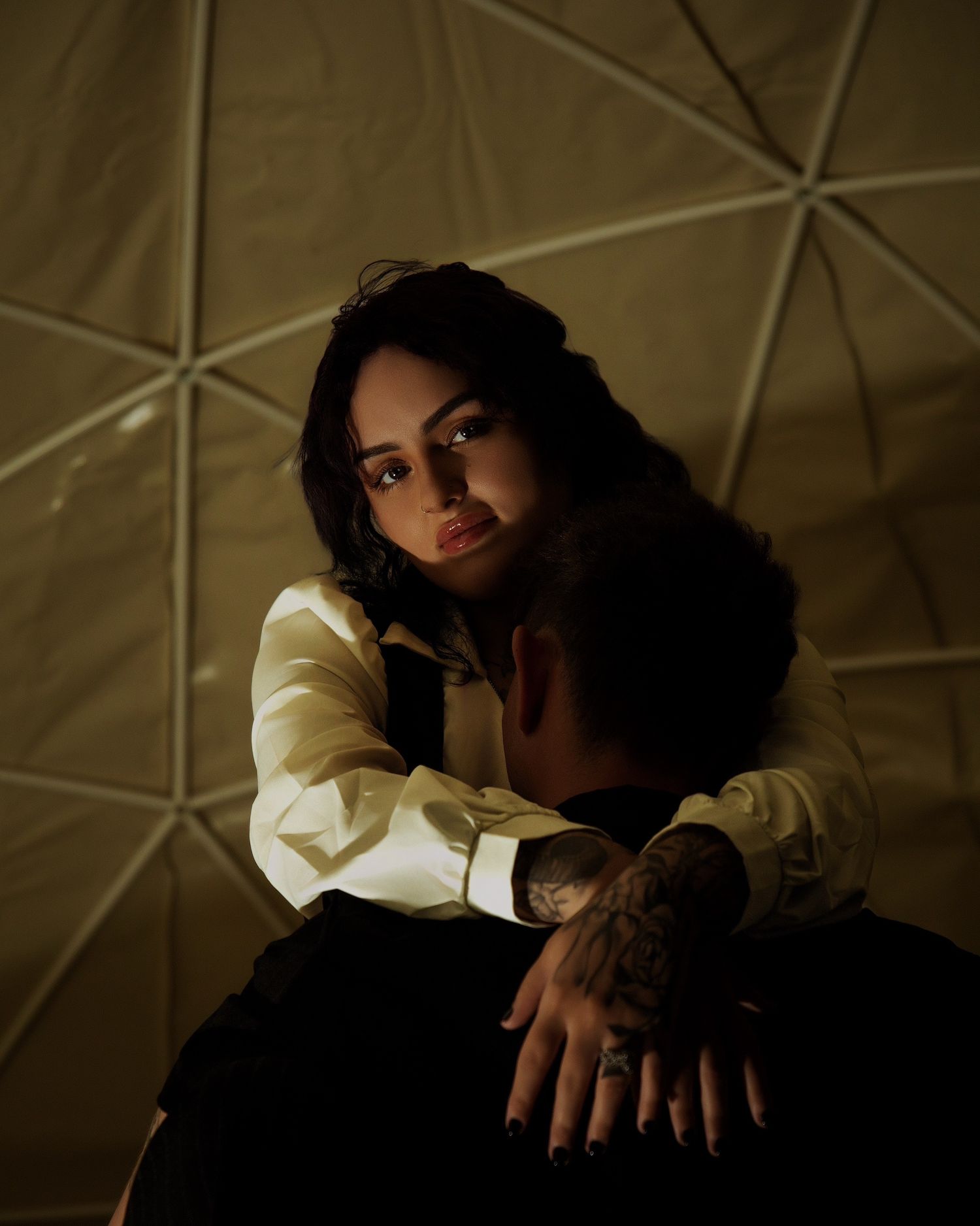 A woman in a white shirt embraces a tattooed man inside a geometric dome, gazing upward in moody lighting.