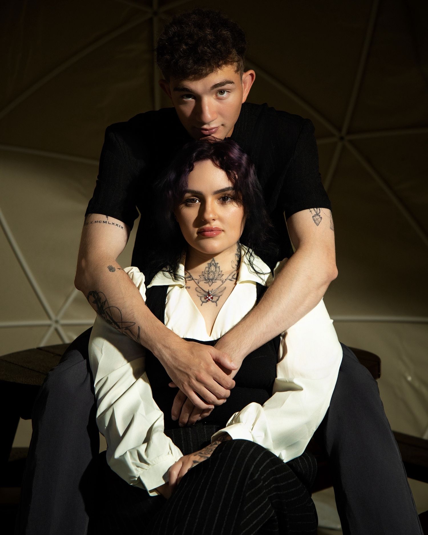 A young couple poses intimately, woman with purple hair and neck tattoo in white blazer, man in black shirt embracing her from behind.