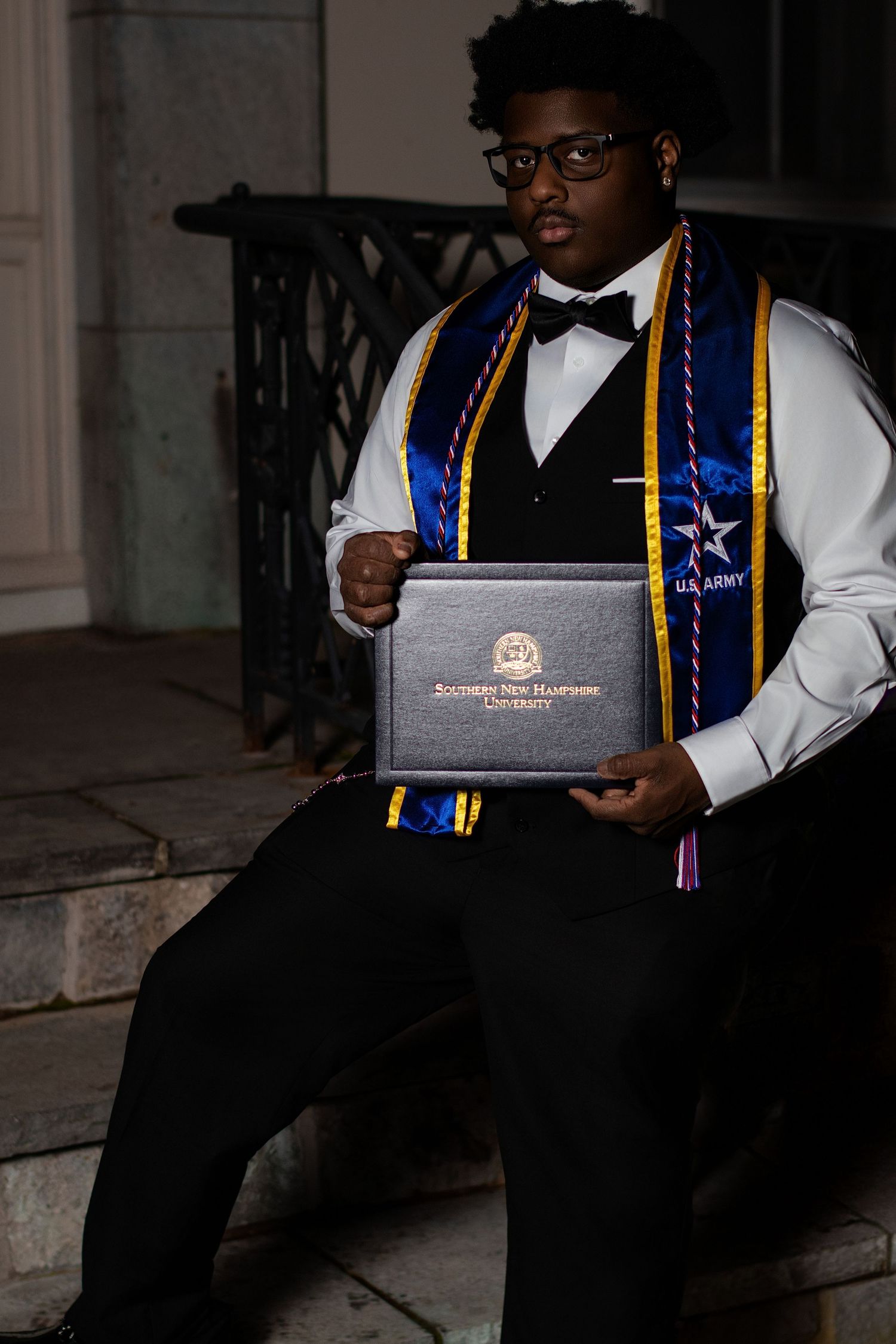 A proud graduate in cap and gown holds a Southern New Hampshire University diploma while wearing a U.S. Army stole.