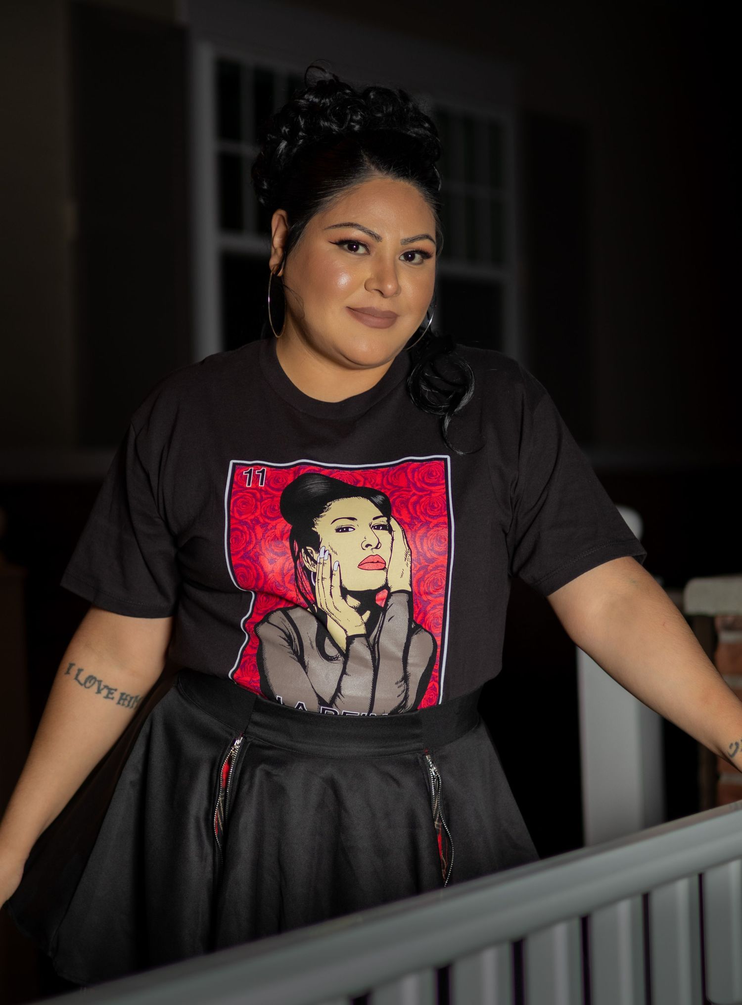 Woman in black graphic tee and skirt poses on porch at night, wearing hoop earrings and hair up.