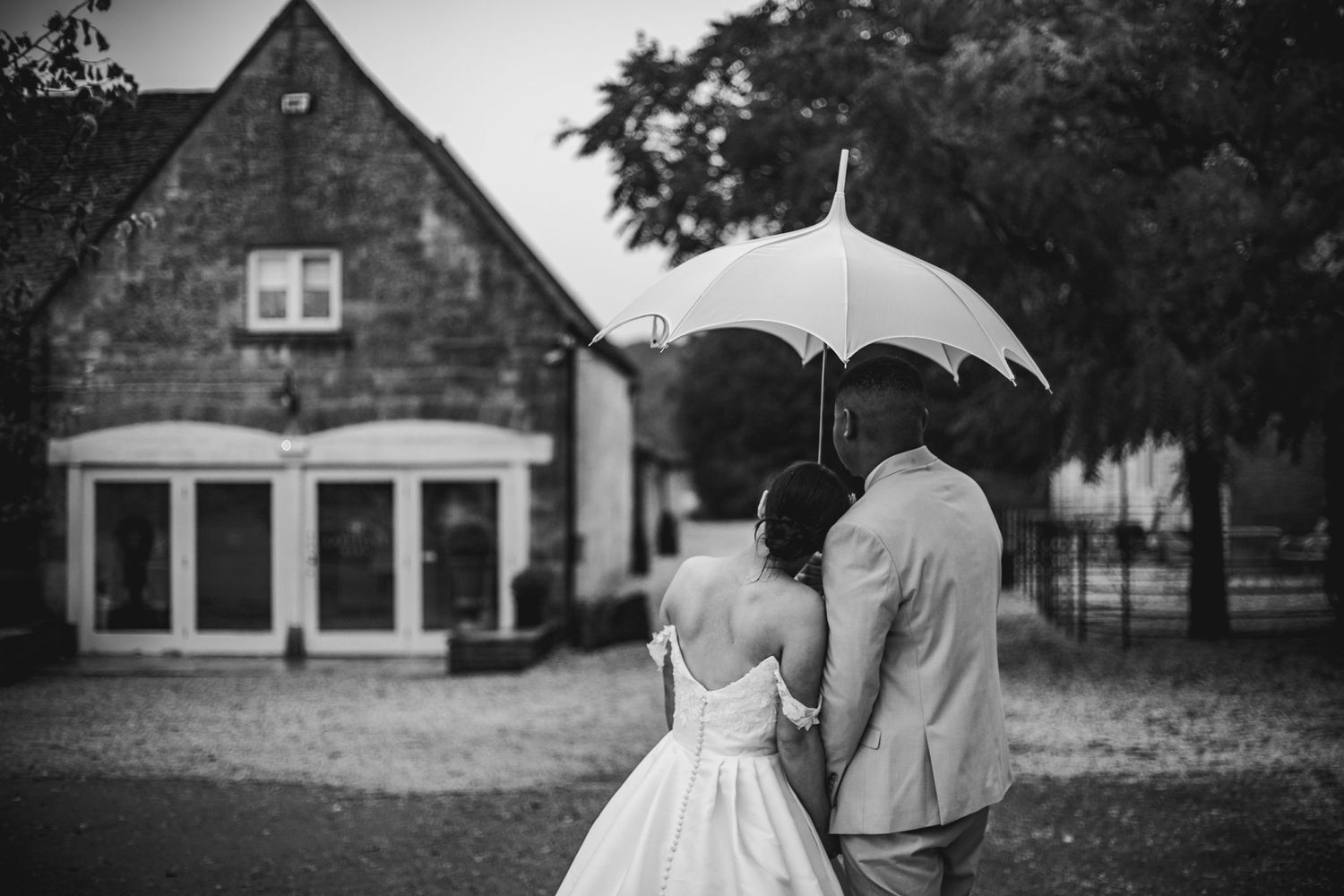 Bride resting her head on groom under a white umbrella outside their wedding venue Lapstone Barn – Chipping Campden, The Cotswolds