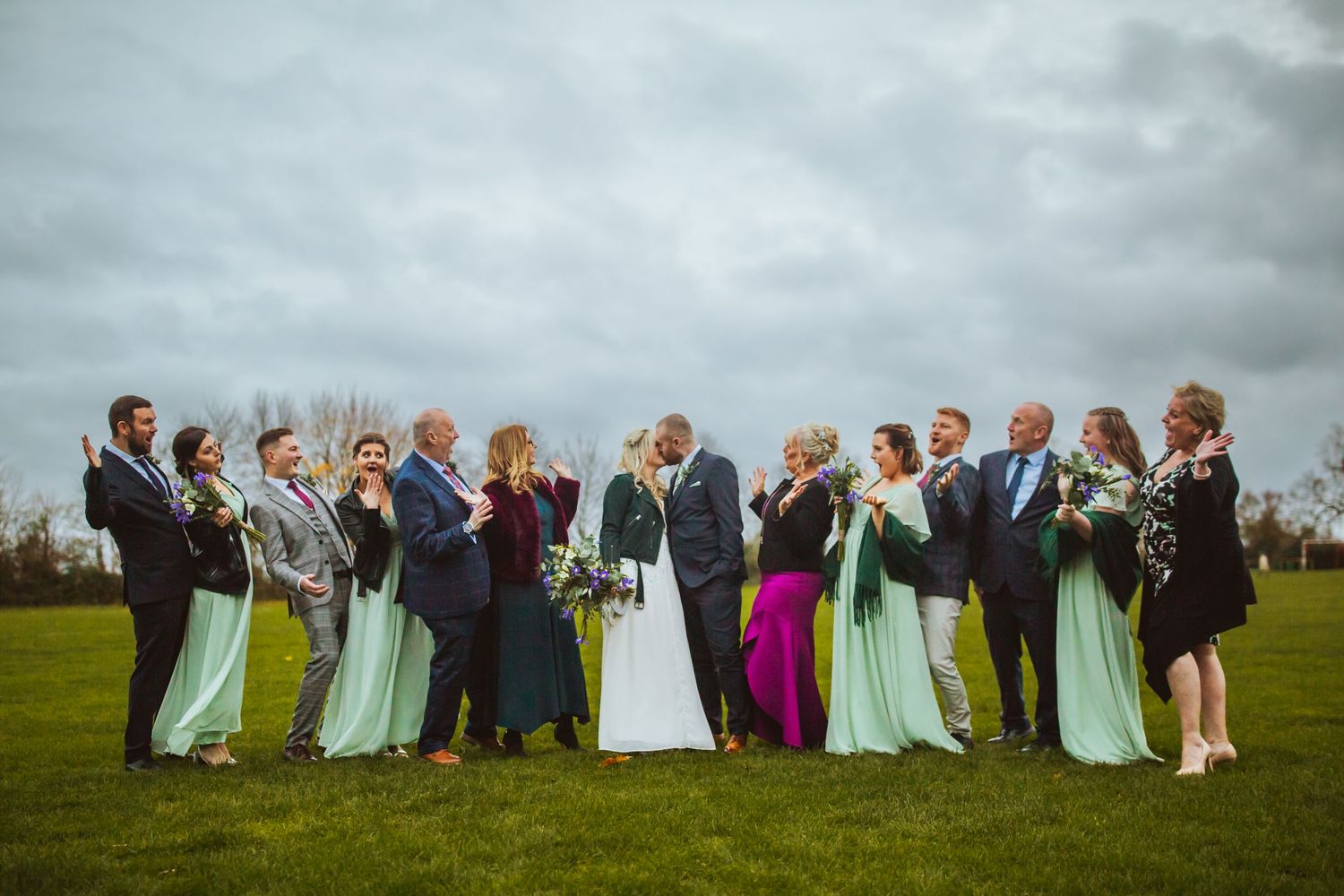 Bride and Grooms family fun pose outside Cottered Village Hall on a field  Hertfordshire, Photographer RELM Photography