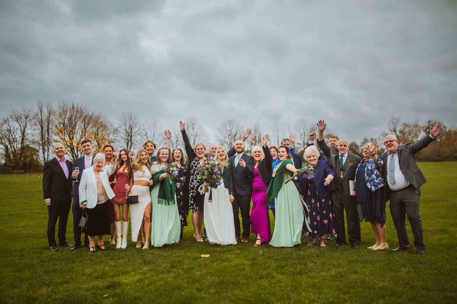 Bride and Grooms family fun pose outside Cottered Village Hall on a field  Hertfordshire, Photographer RELM Photography