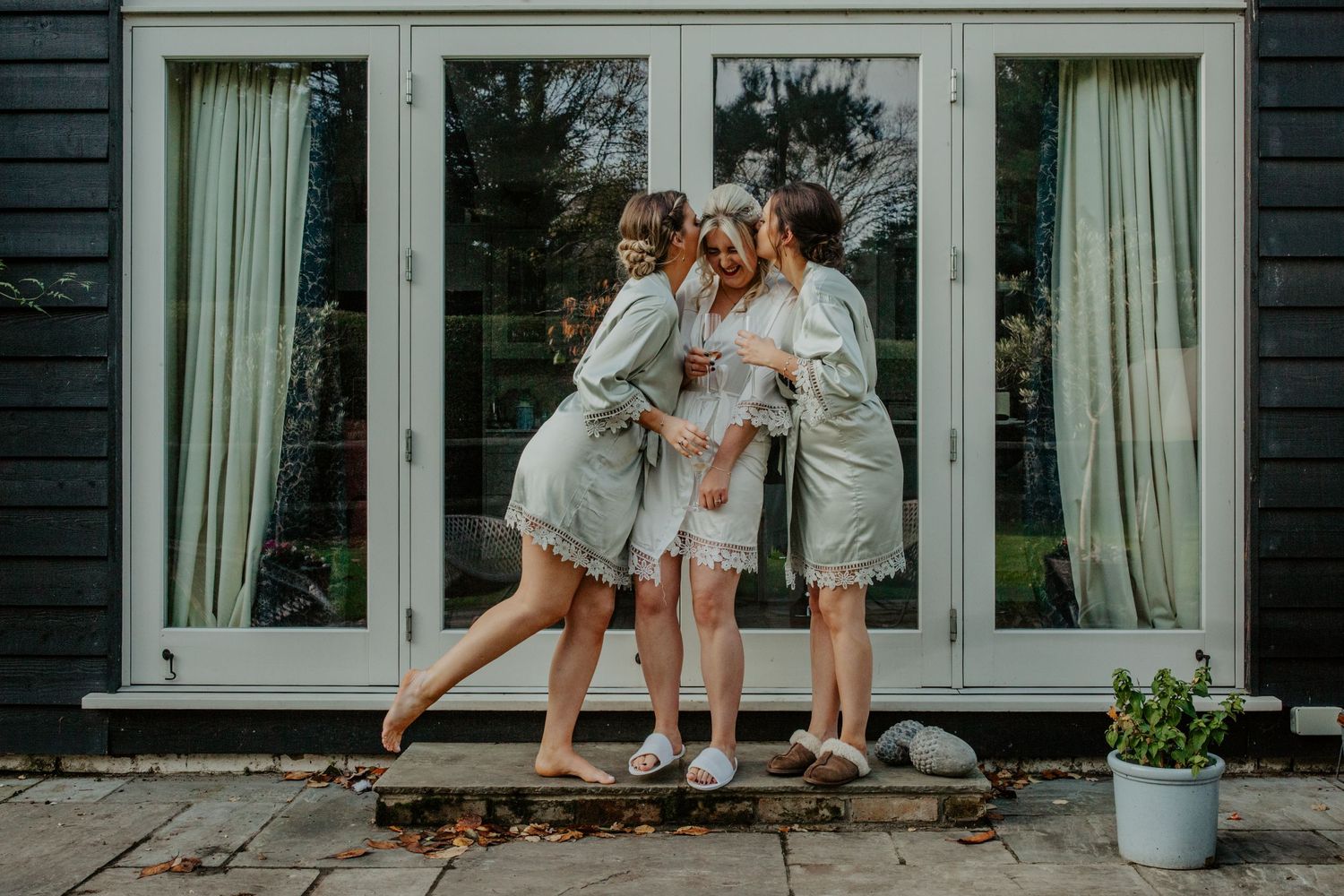 Bride being kissed on the cheeks by her sisters in their bridal robes in Hertfordshire, Photographer RELM