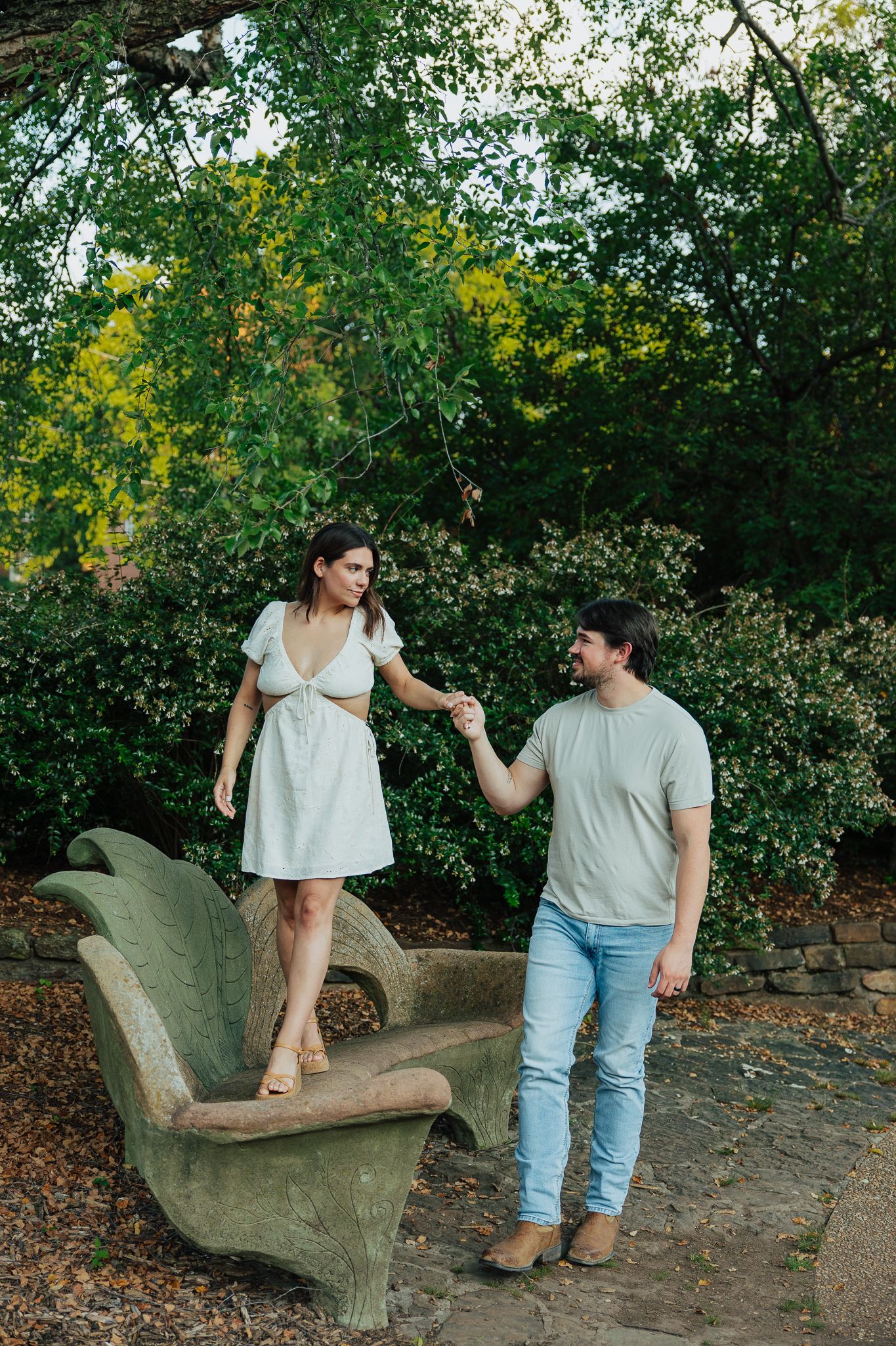 Couple holding hands in a lush green garden, woman standing on a decorative stone bench.