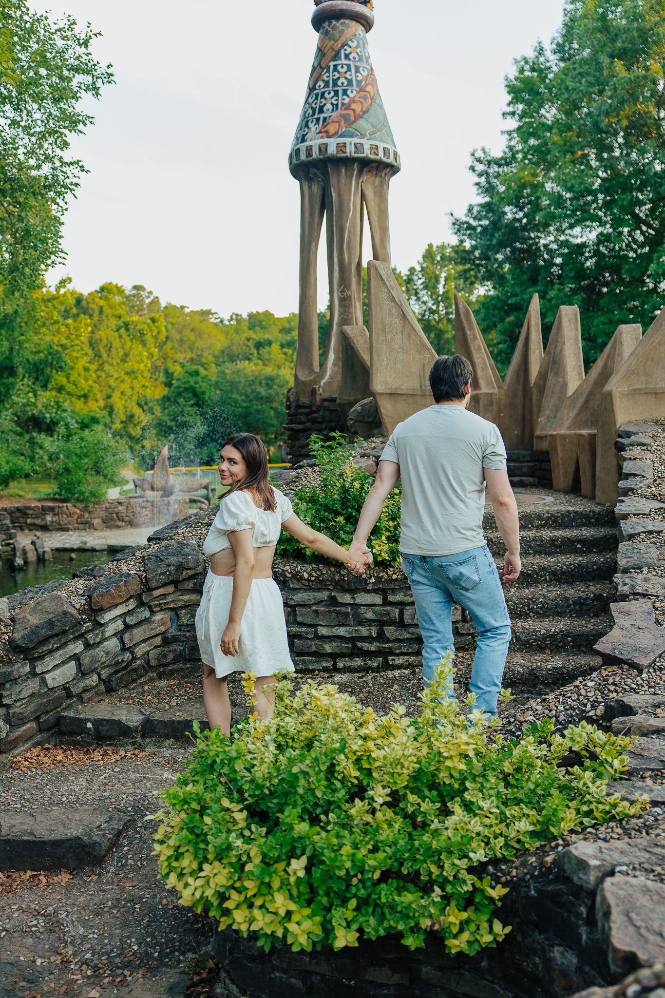 Couple holding hands walking up stone steps toward ornate mosaic tower sculpture in lush garden park.