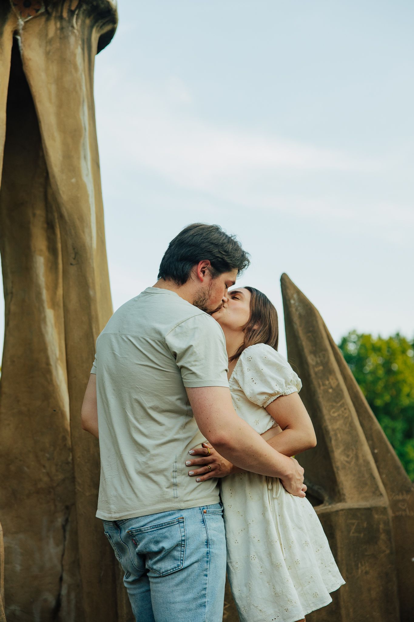 Couple sharing a romantic kiss between large stone sculptures in a scenic outdoor garden setting.
