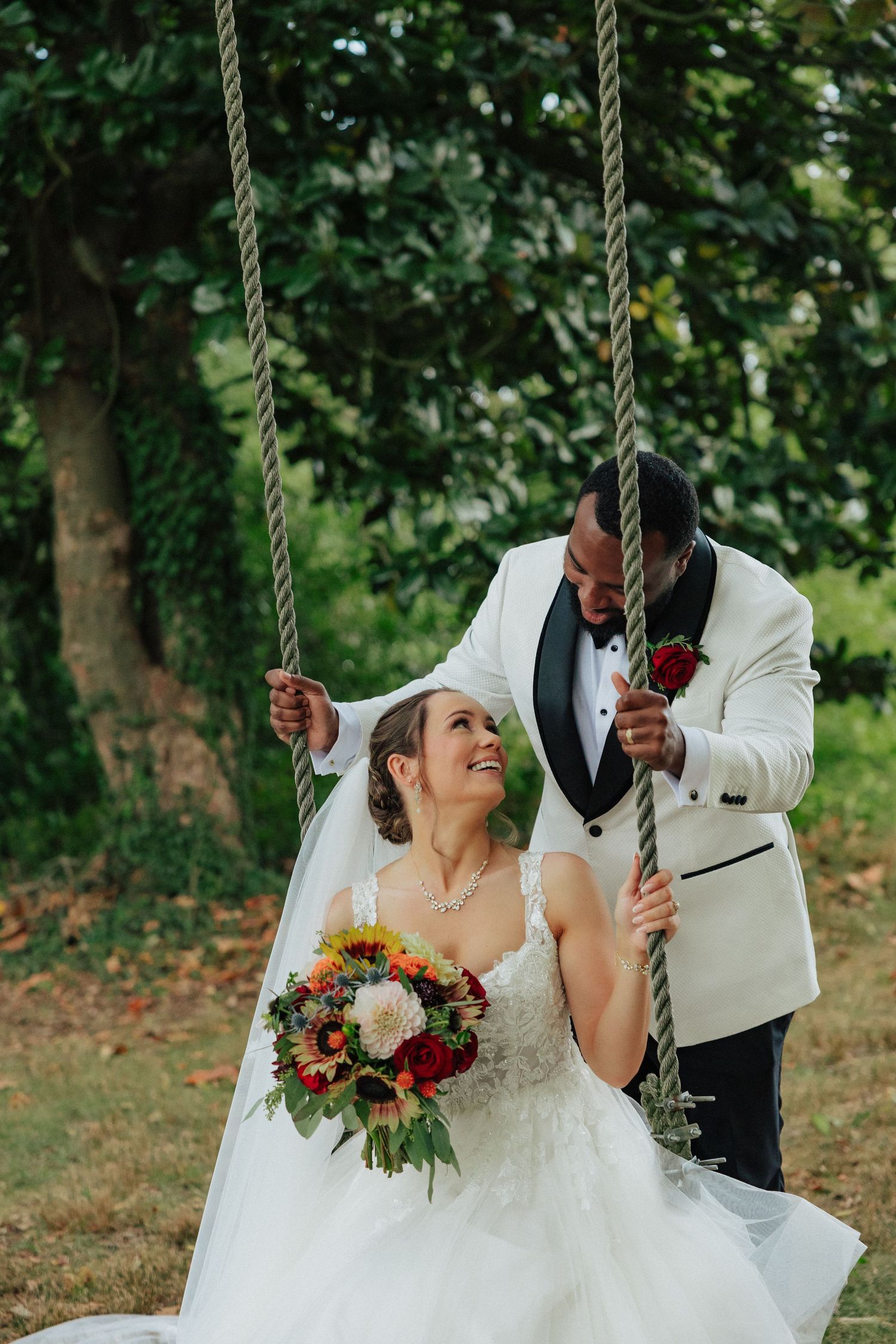 Groom in white tuxedo pushes bride on rope swing as she laughs holding colorful bridal bouquet outdoors.