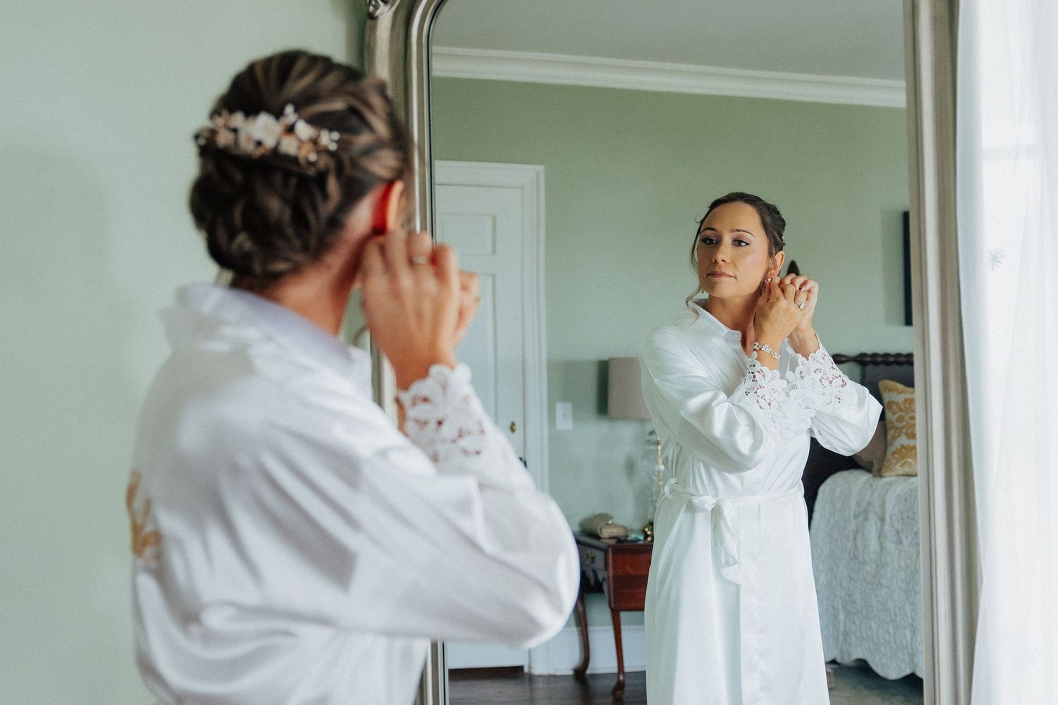 Bride in white robe adjusts earrings while looking in mirror during wedding morning preparation routine.