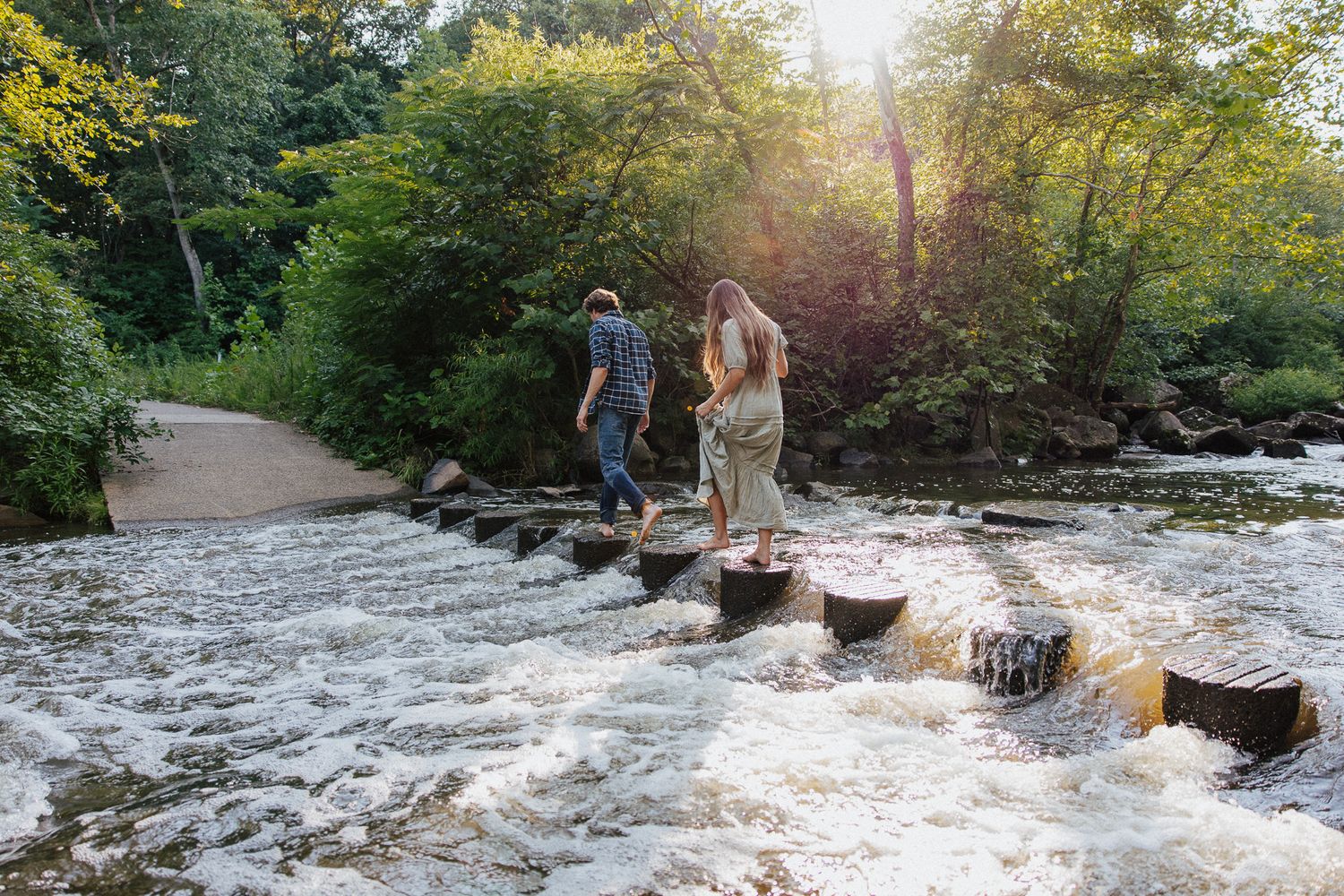 A couple carefully steps across stone river crossings surrounded by lush green trees in soft sunlight.