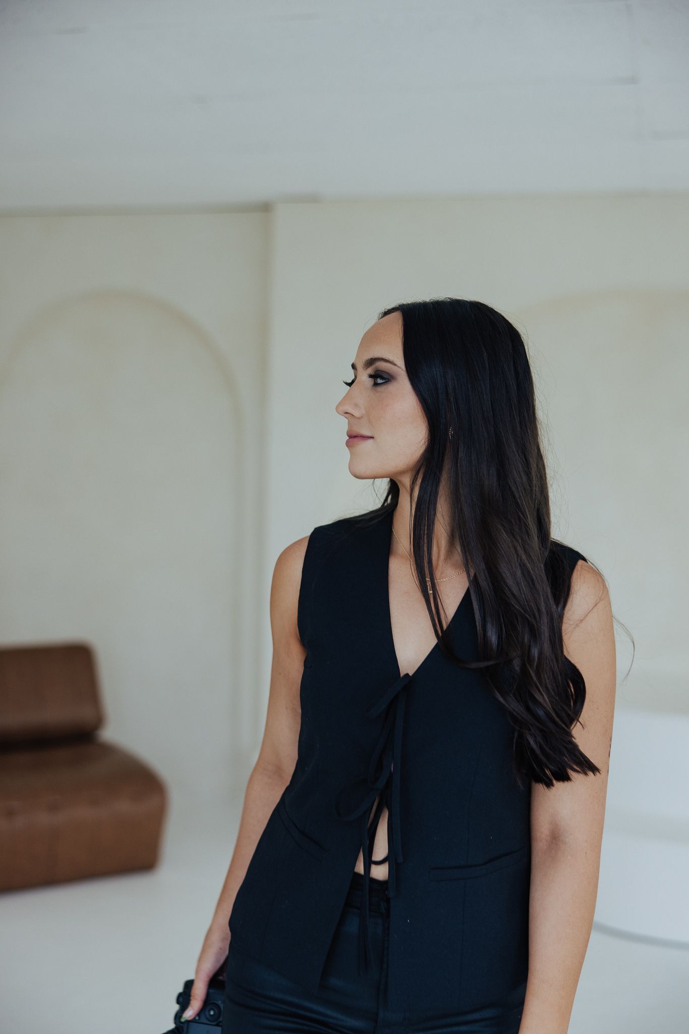 Woman with long dark hair wearing a sleeveless black tie-front outfit standing in a bright minimalist room.