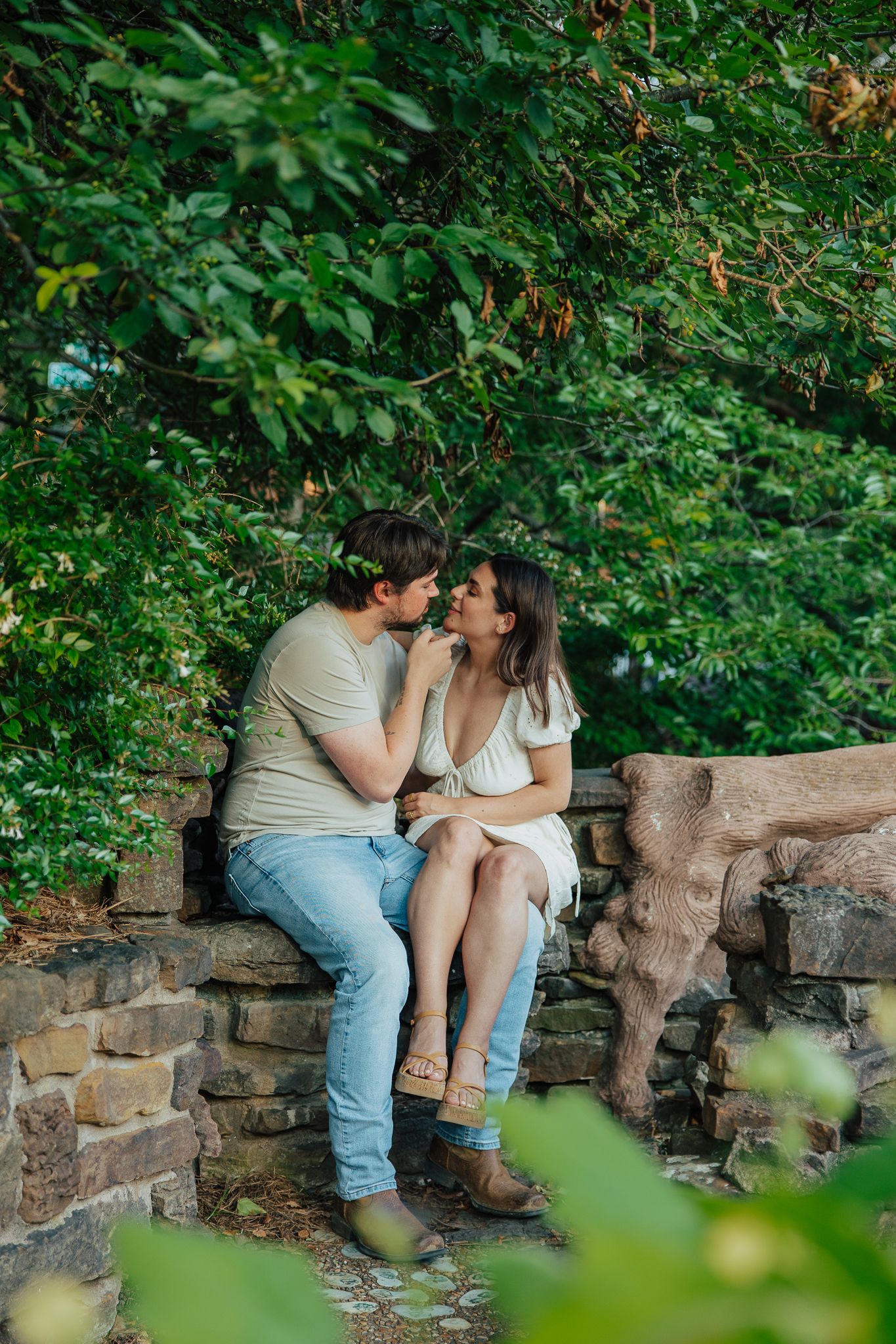 Couple sharing a romantic moment sitting on a stone wall surrounded by lush green trees in a garden.