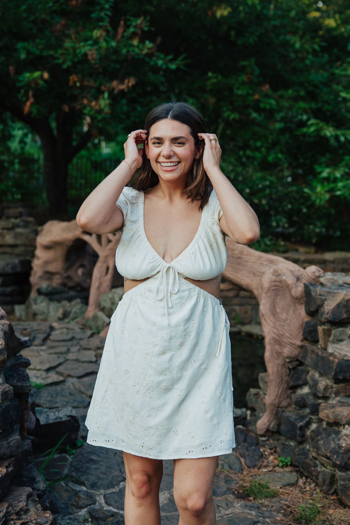 A happy woman in a white cut-out dress laughs while adjusting her hair in a garden setting.