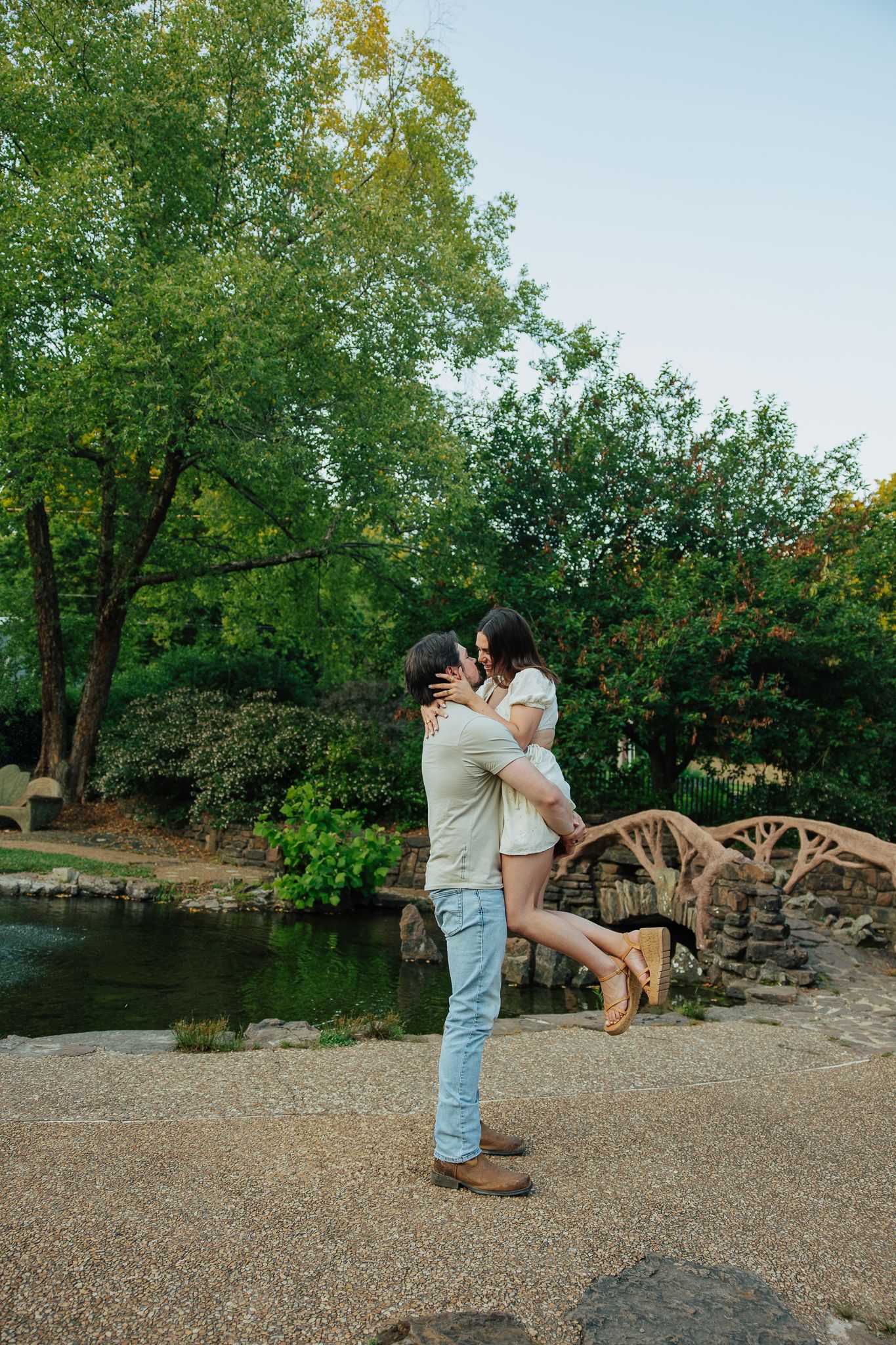 Man lifting woman for a kiss near scenic pond with wooden arched bridge in lush green park.