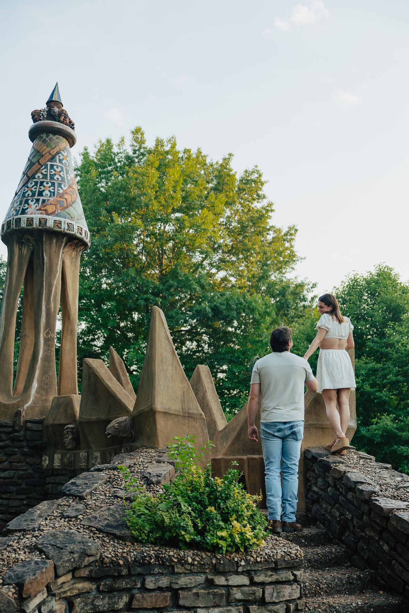 Romantic couple holding hands standing atop stone wall with decorative mosaic spire sculpture outdoors.
