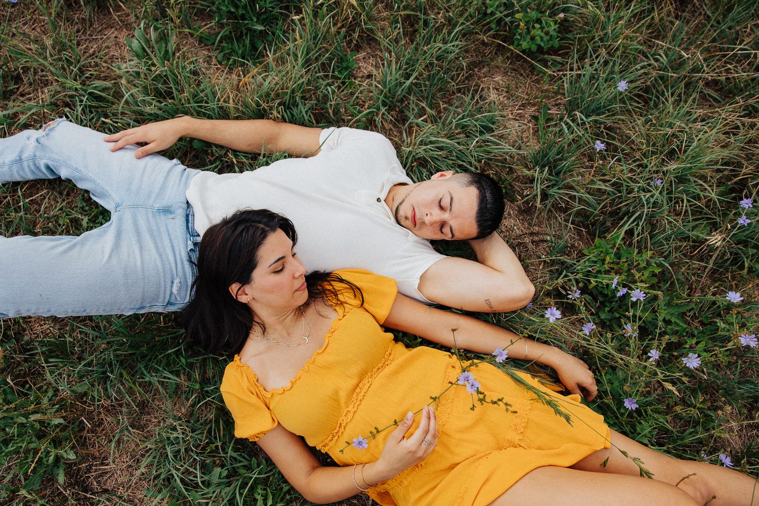 A couple lying in a grassy field with wildflowers, woman in yellow dress resting her head on mans shoulder, eyes closed.