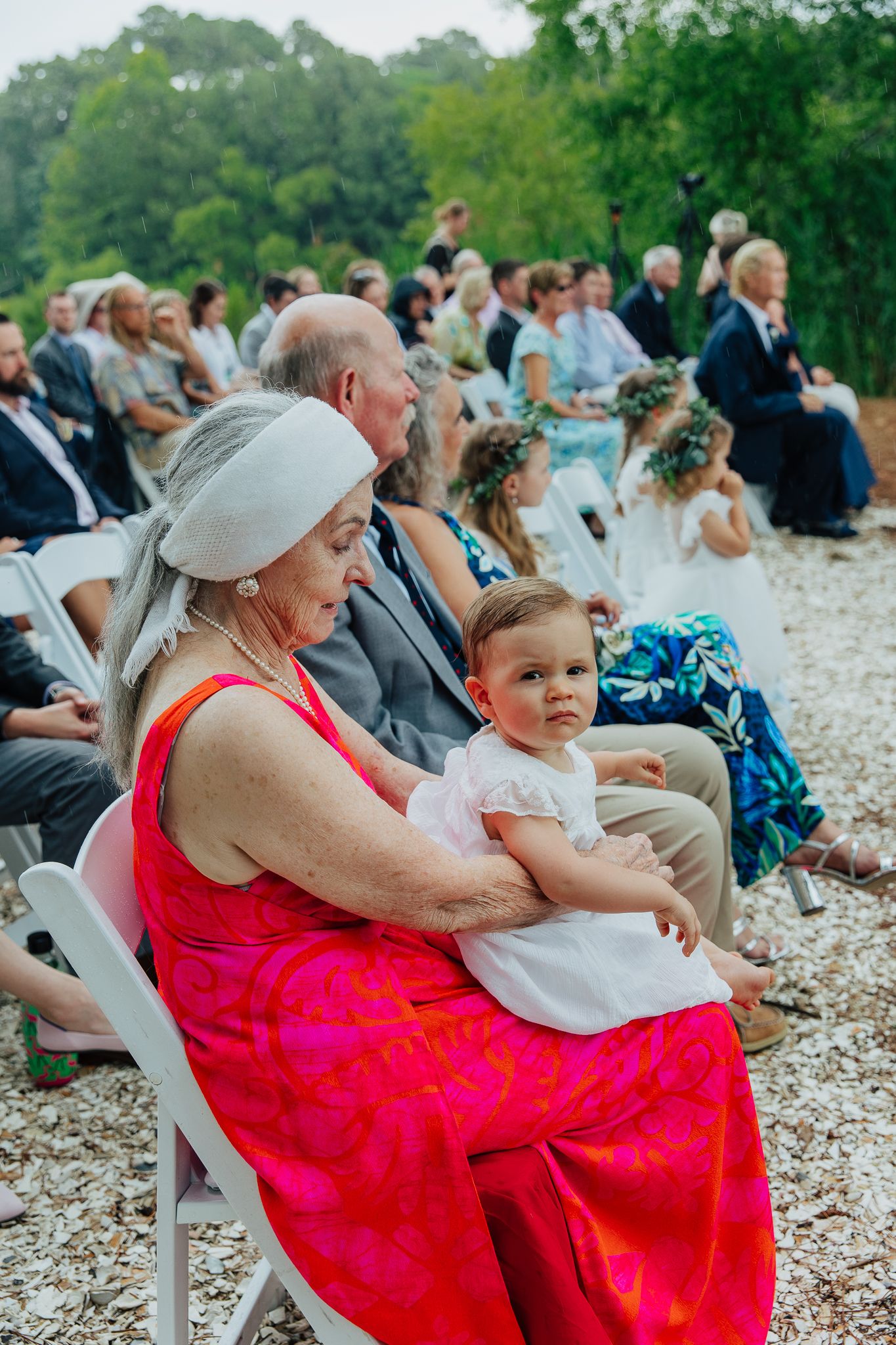 Elderly woman in red dress holds baby at outdoor wedding ceremony with guests seated on white chairs.