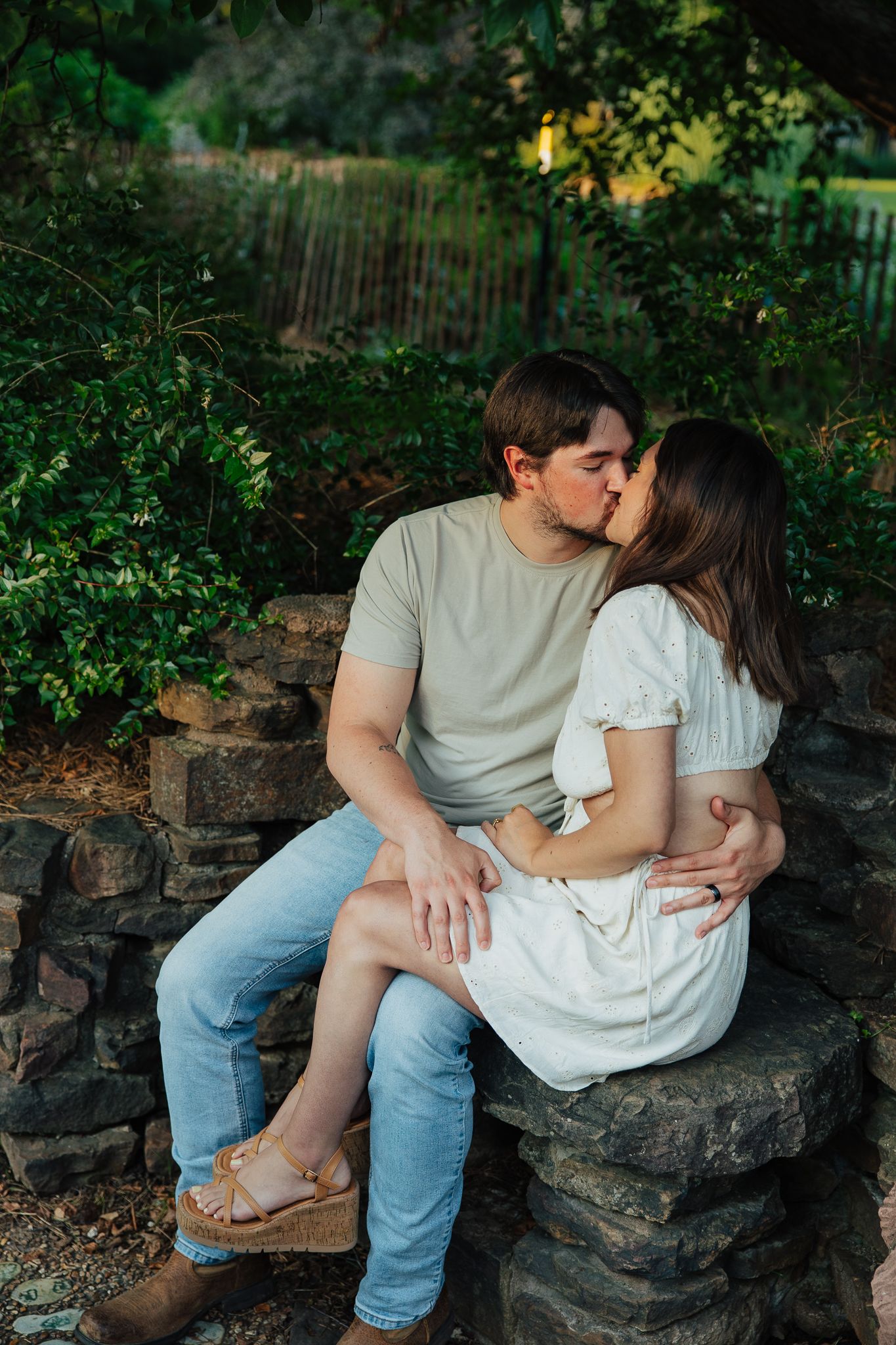 Couple sharing a romantic kiss while seated on stone wall in sunlit garden.