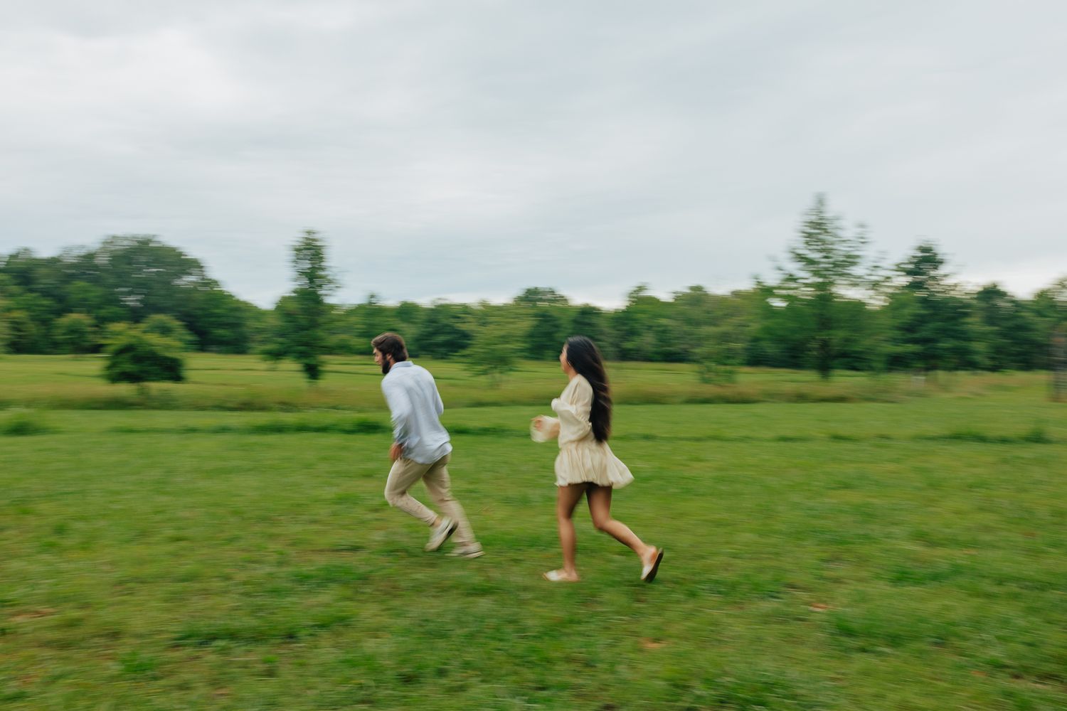 Couple runs playfully across vast green meadow on overcast day during outdoor engagement photography session.