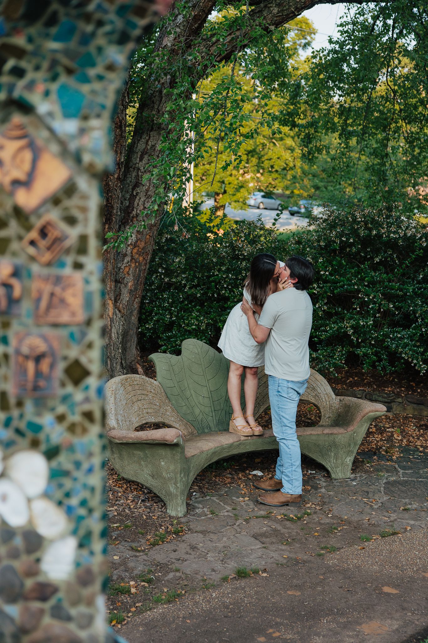Couple sharing a kiss on a decorative stone bench in a garden framed by a mosaic archway.