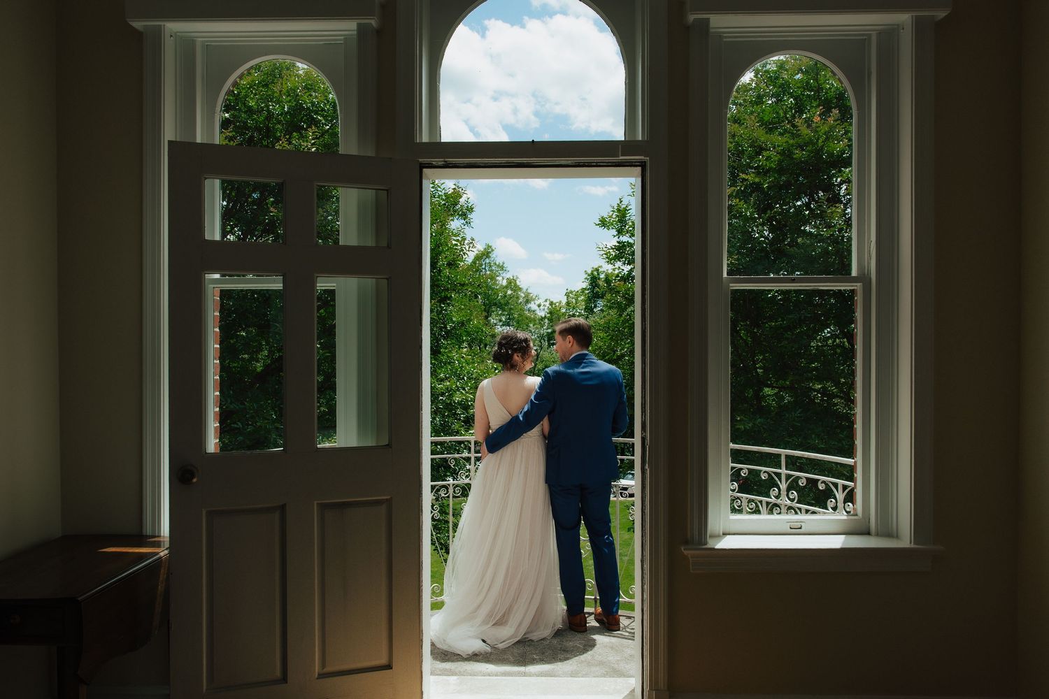 Bride and groom stand in elegant arched doorway overlooking lush green garden on their wedding day.