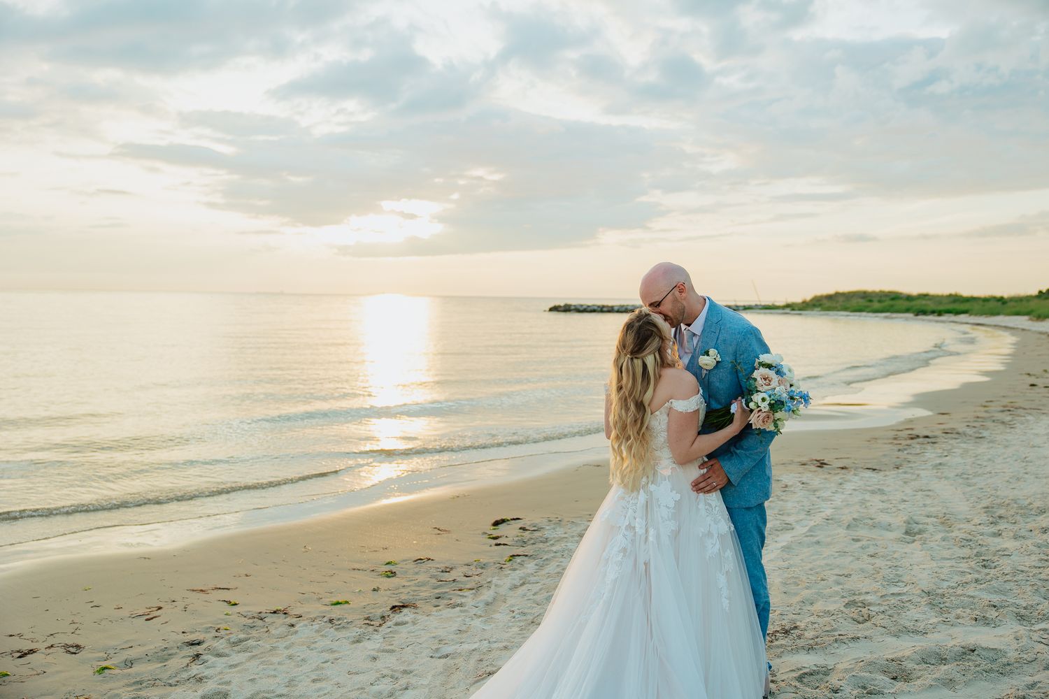 Newlyweds share a kiss on a scenic beach at golden sunset, groom in blue suit holding floral bouquet.