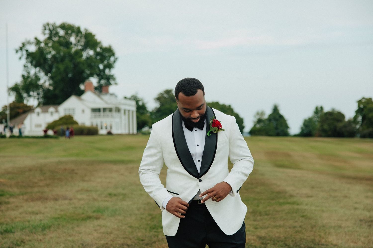 Groom in white tuxedo jacket with red boutonniere adjusts buttons on green lawn at wedding venue.