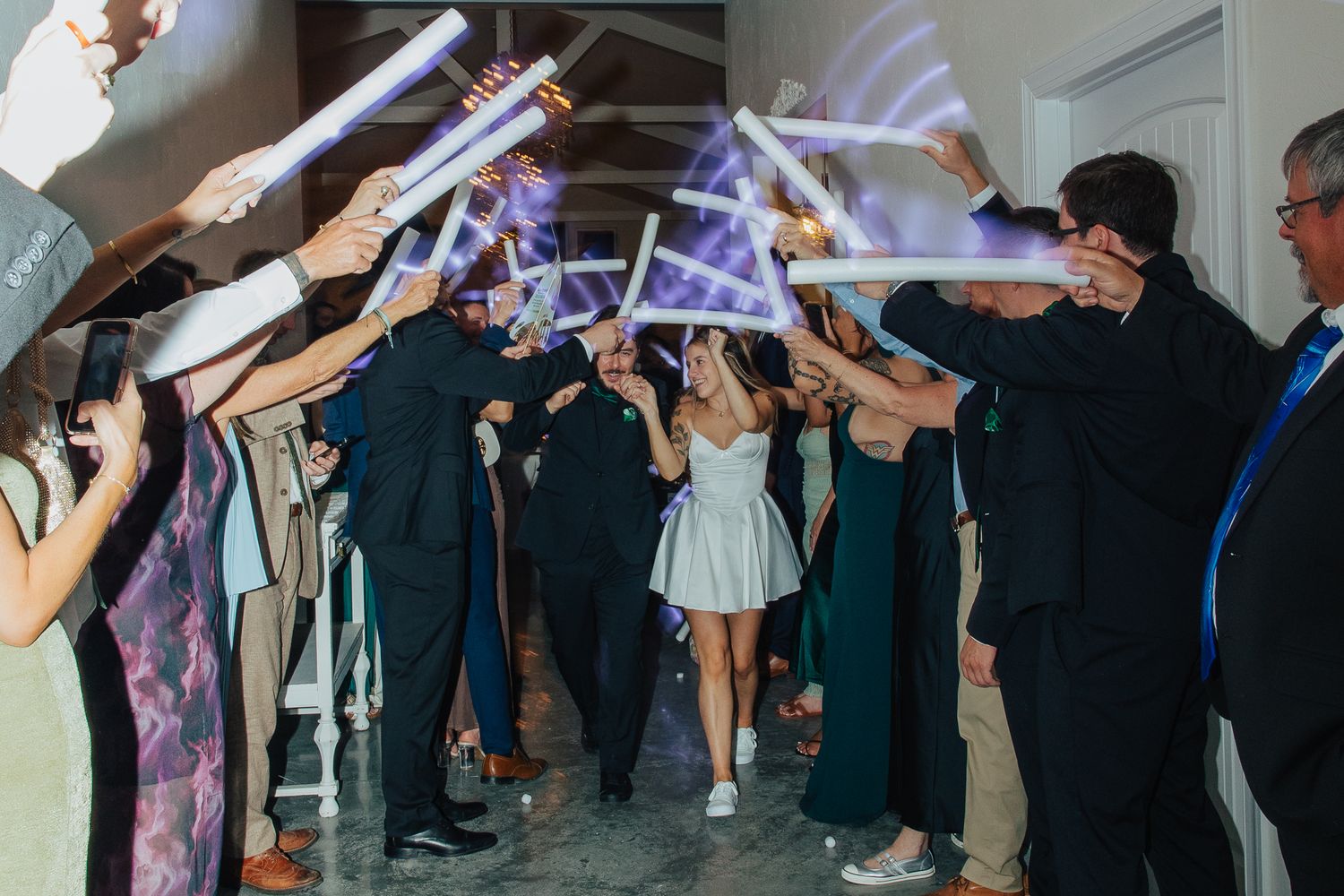 A bride in a short white dress walks through a glowing light saber send-off at her wedding reception.