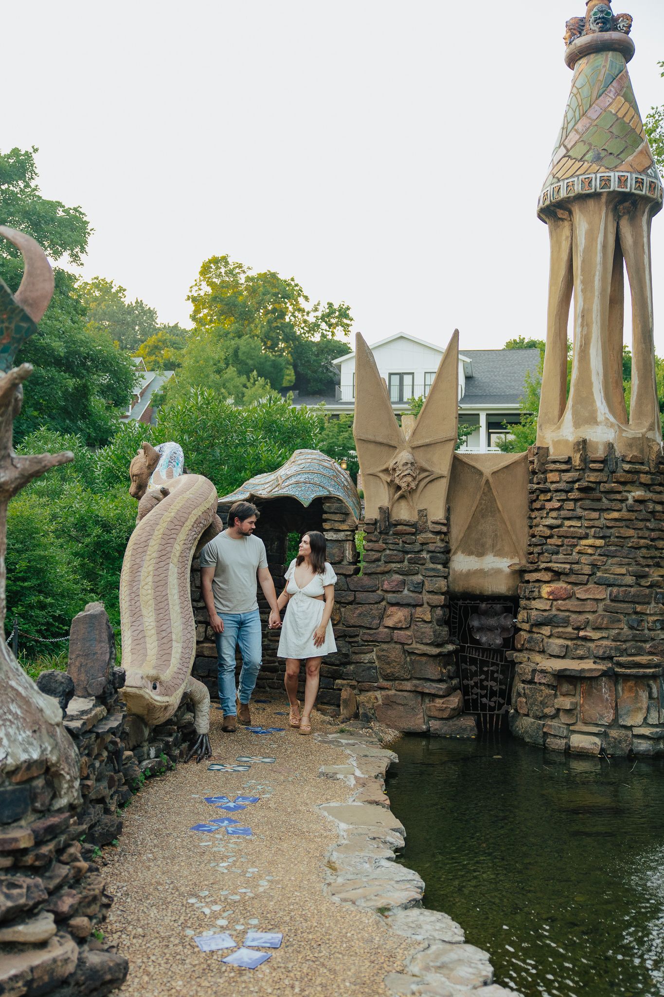 Couple walking hand in hand through whimsical sculpture garden with mosaic tower and stone archway.
