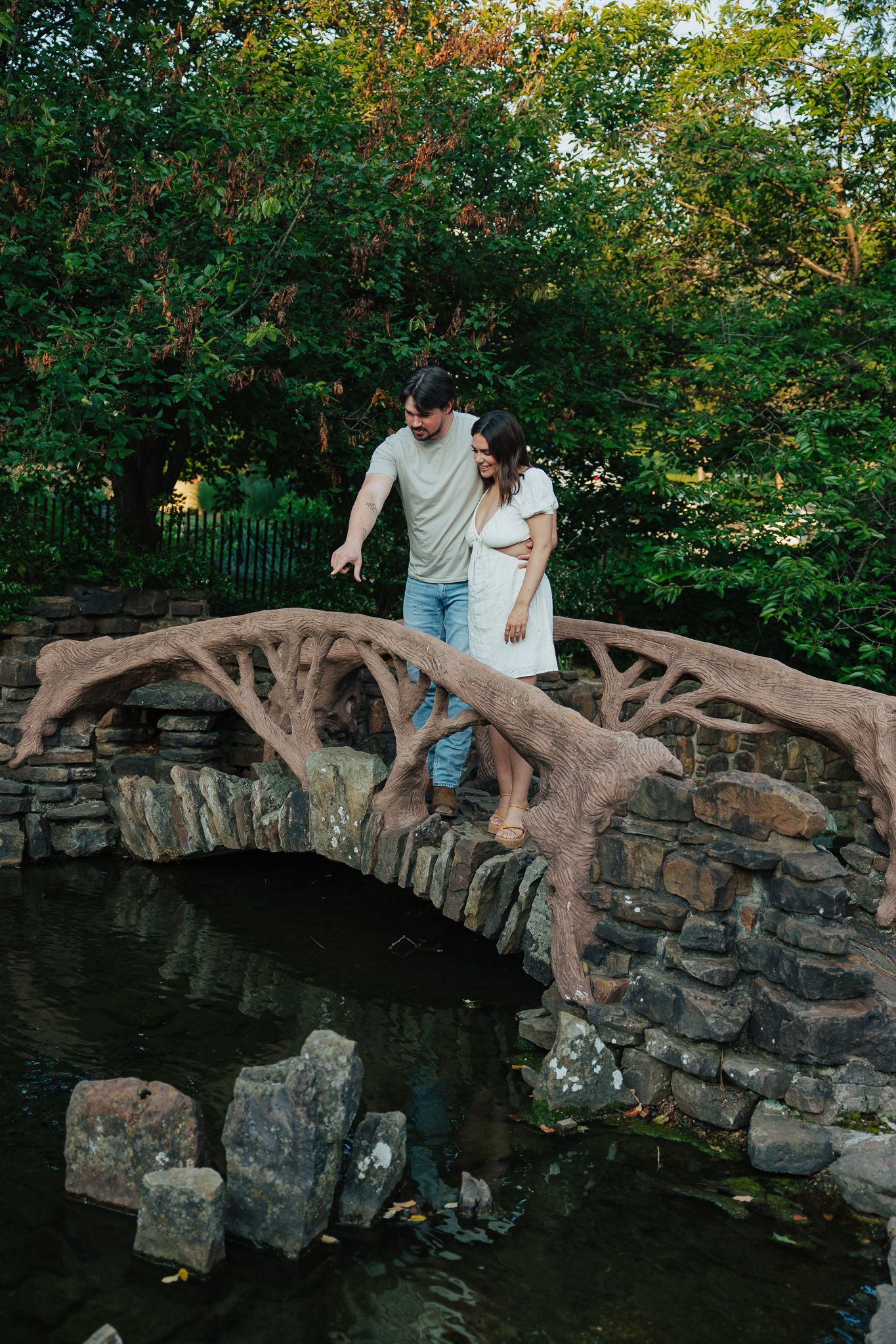 Couple walking hand in hand on ornate stone arch bridge over garden pond.