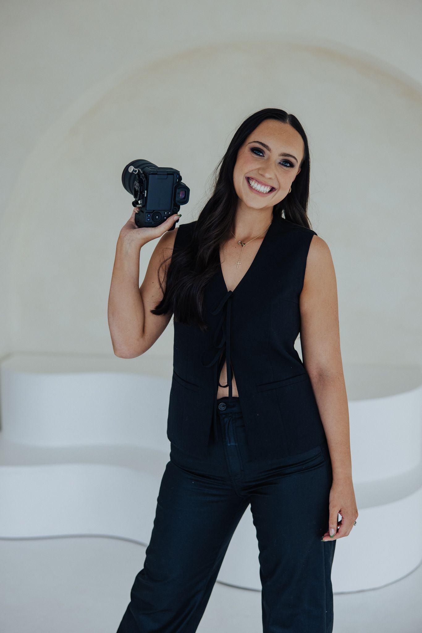 A smiling female photographer in all-black outfit holds a DSLR camera up in a modern white studio.