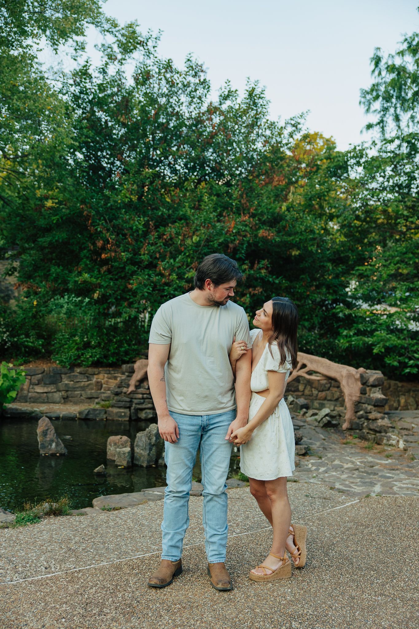 Couple standing close together holding hands near a peaceful pond with stone bridge in park.