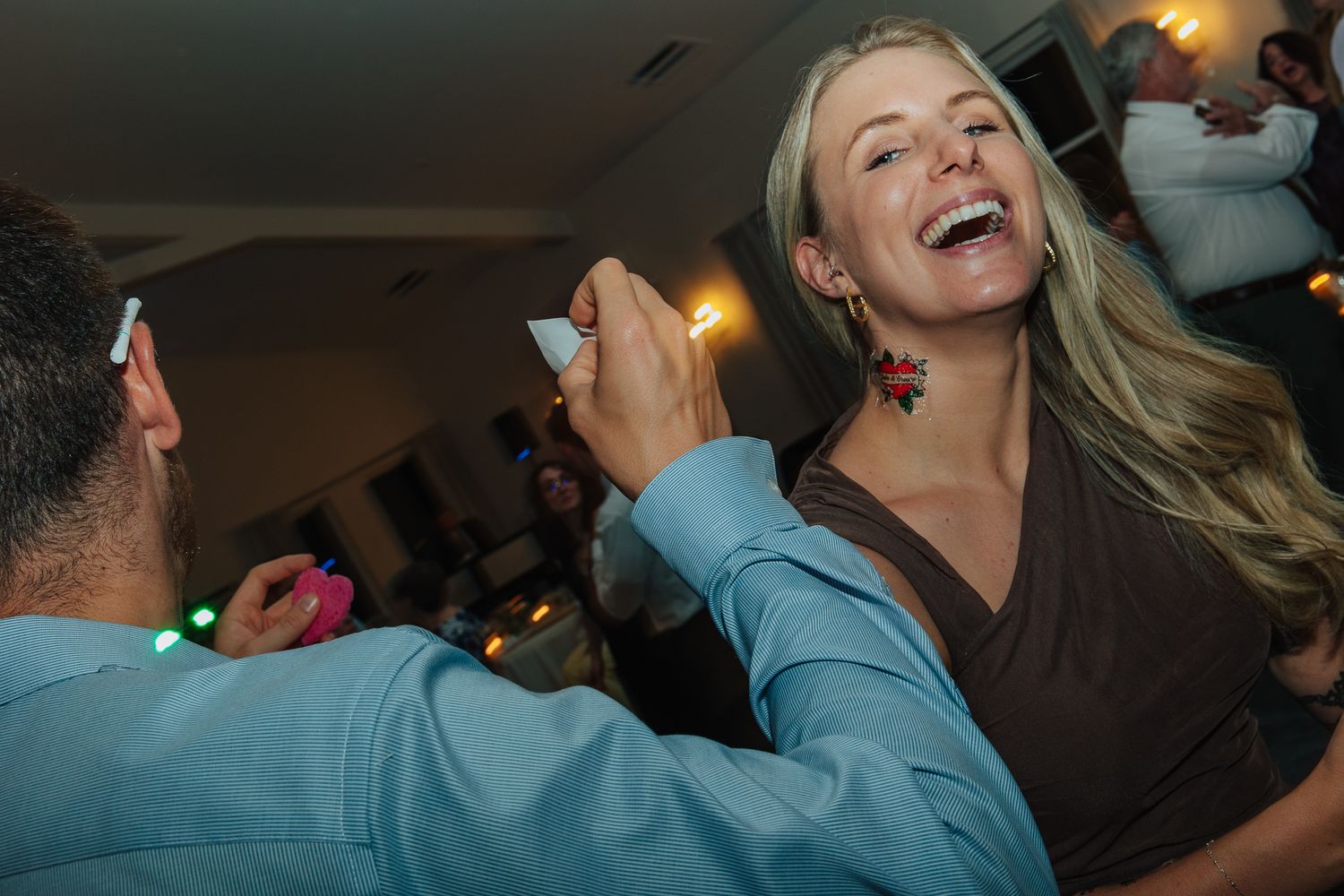 A laughing blonde woman in a brown dress dances with a man in a blue shirt at a wedding reception.