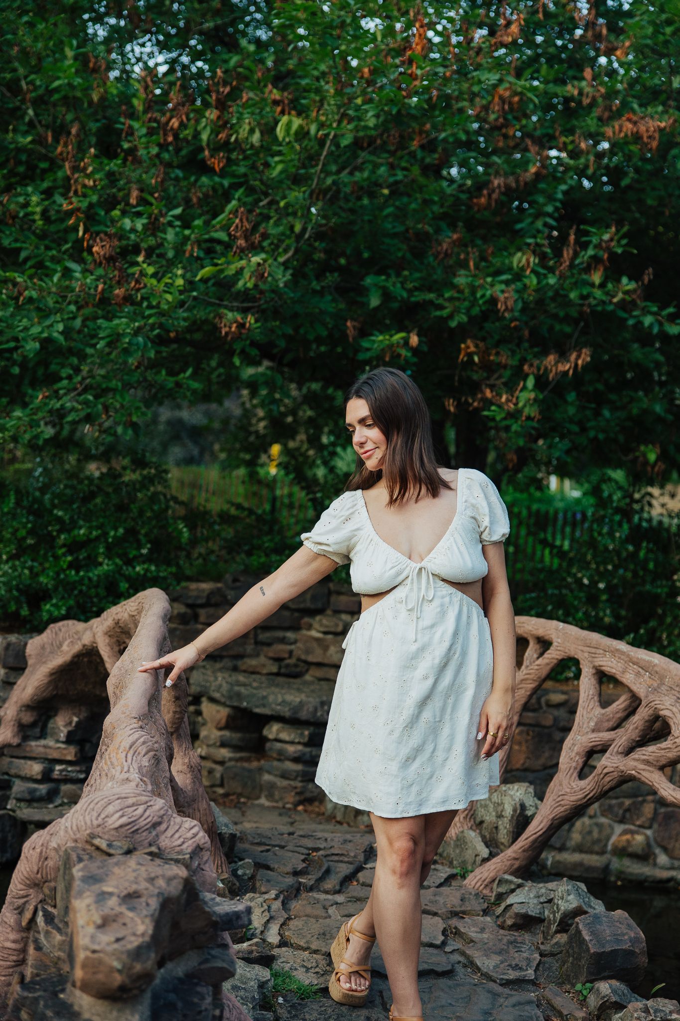 A smiling woman in a white eyelet mini dress poses on a rustic stone bridge in a garden.