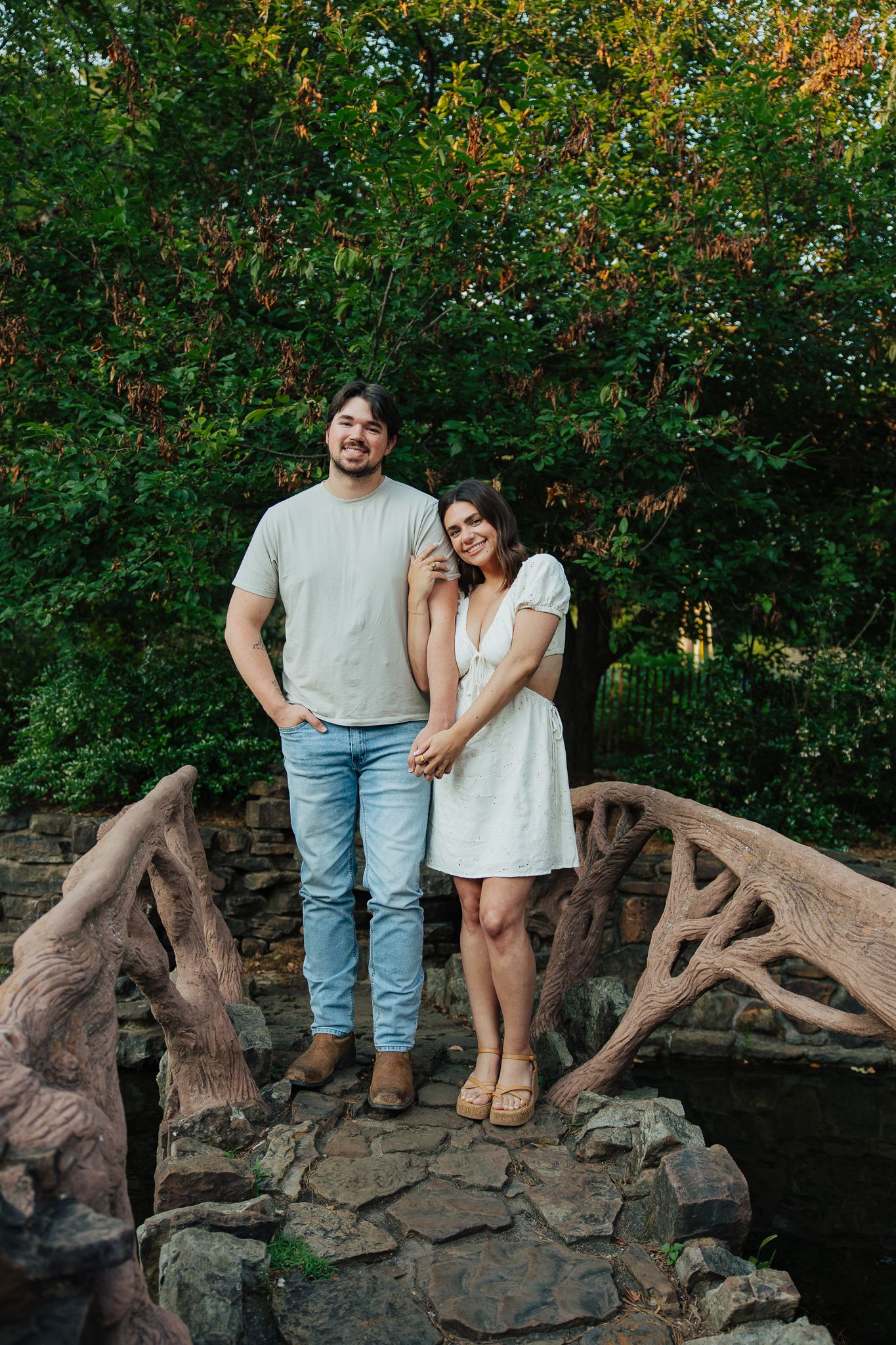 Couple posing together on rustic stone bridge surrounded by lush green garden trees.