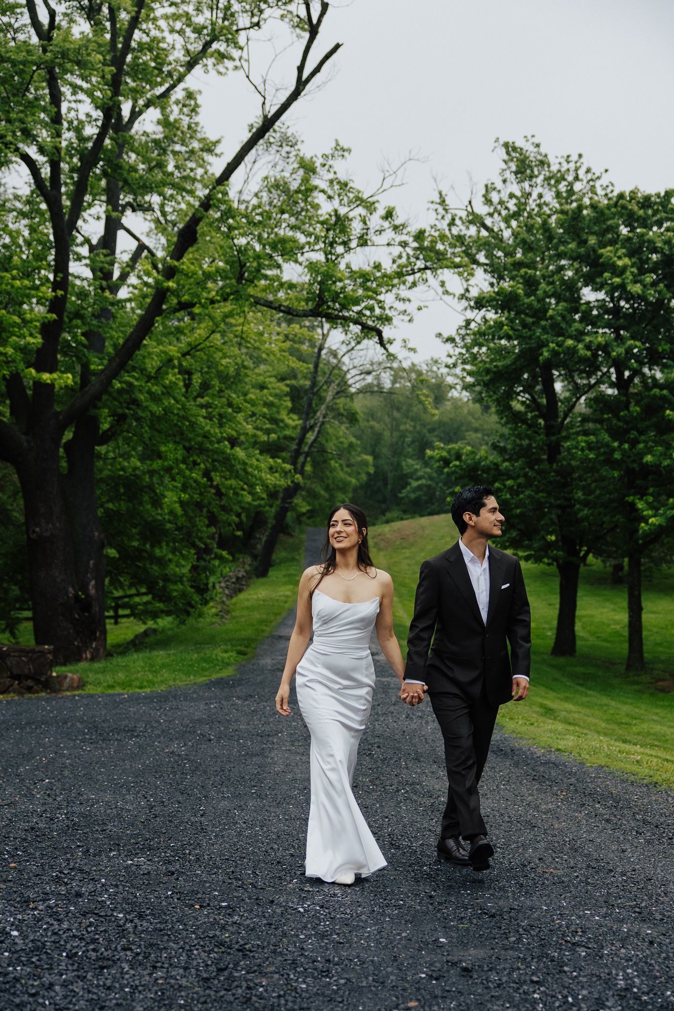 A bride in a white gown and groom in a black suit walk hand-in-hand down a tree-lined gravel path.