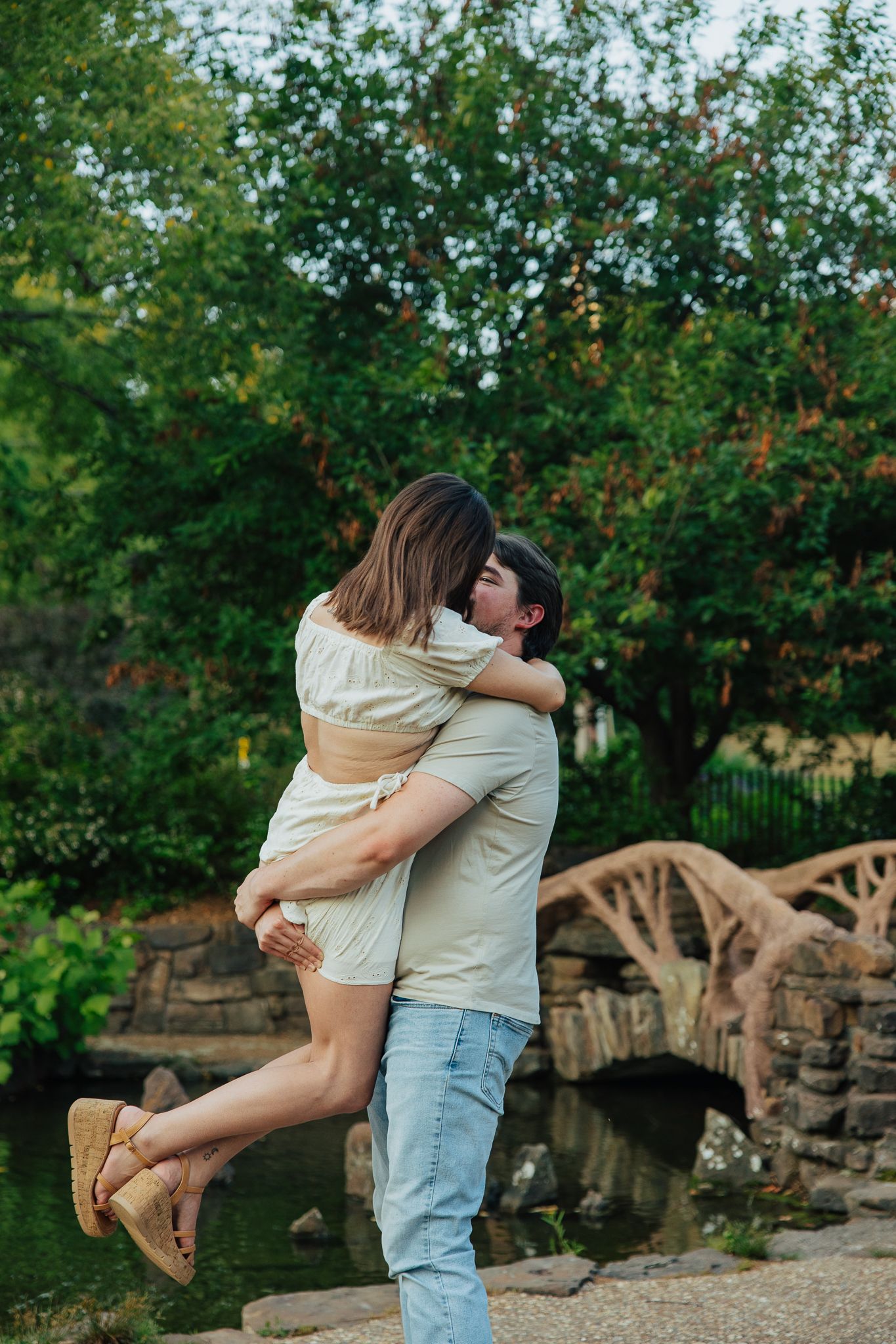 Man lifting and kissing woman near a tranquil pond with wooden bridge in a green garden park.