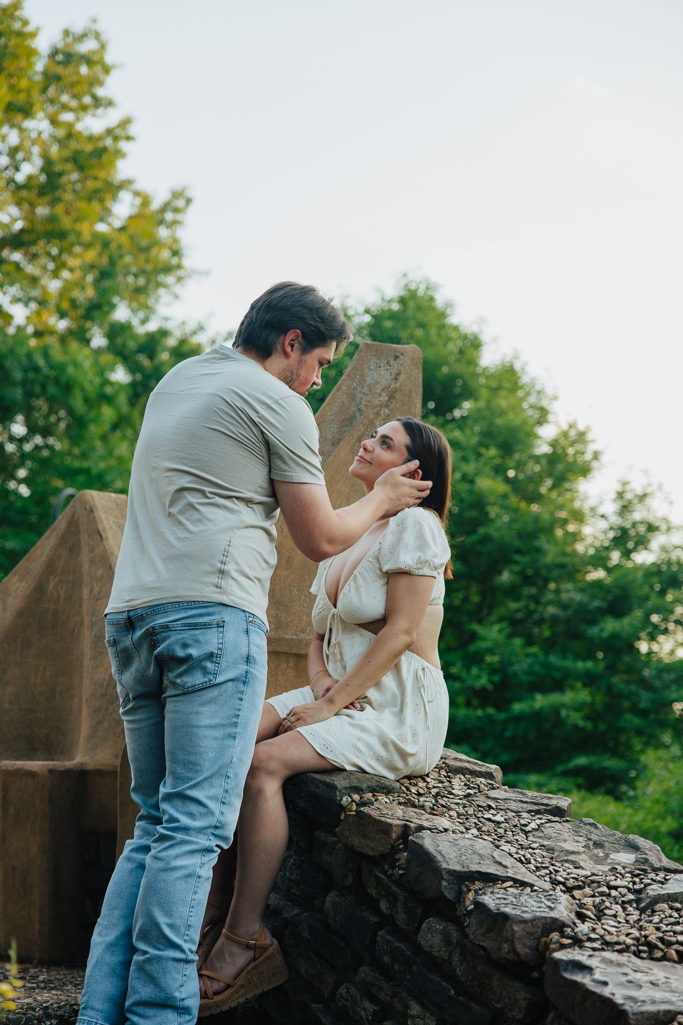 Man tenderly touching womans face as she sits on stone wall surrounded by trees in outdoor park.