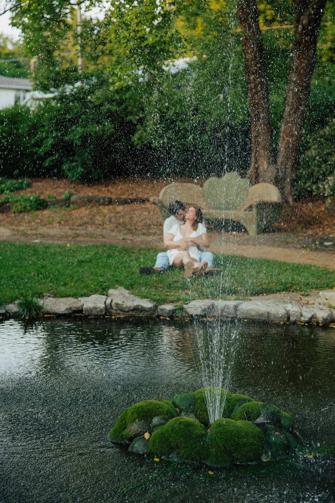 Couple sitting together on grass near a pond with a fountain in a peaceful garden park.
