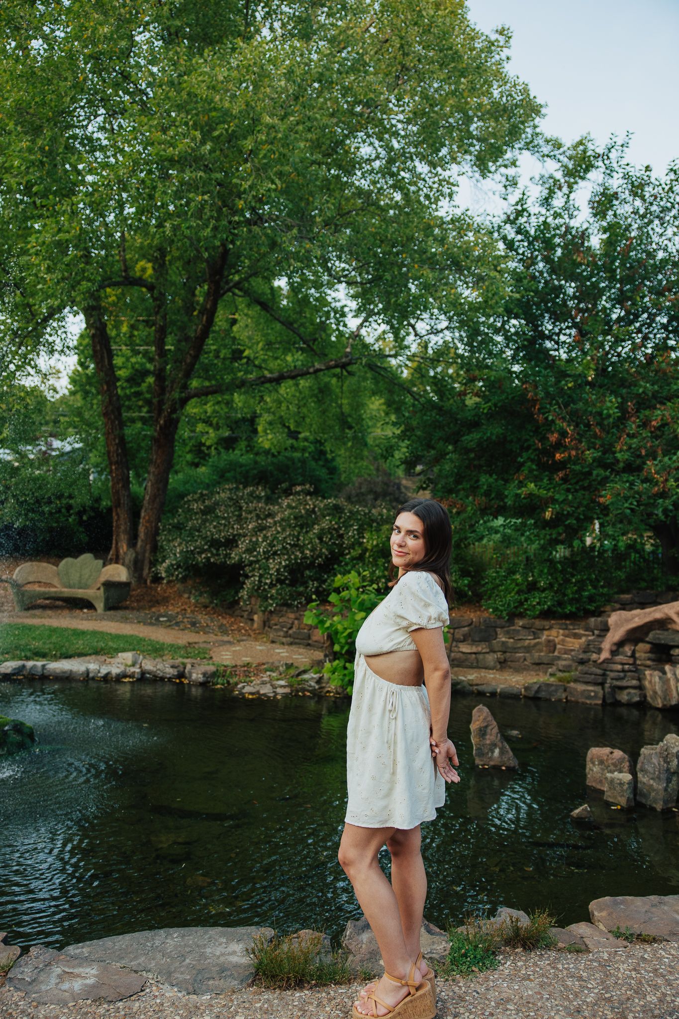A woman in a white mini dress stands by a serene pond surrounded by lush green trees.