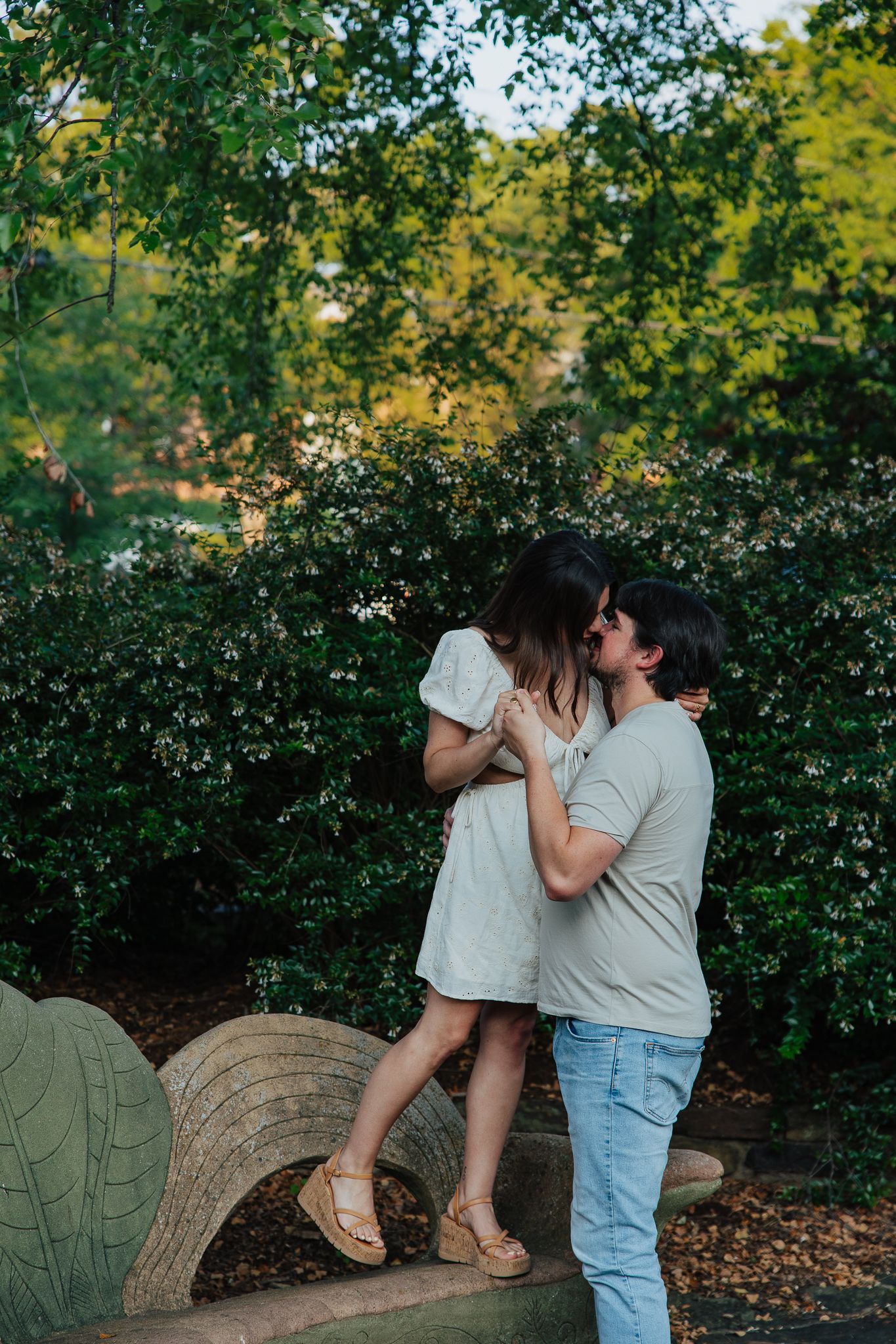 Romantic couple kissing near a decorative stone bench surrounded by blooming white flowers.