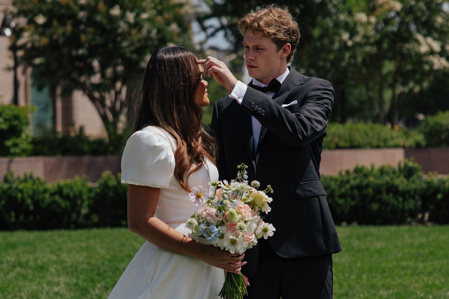 Groom tenderly touches bride's face outdoors, bride holds wildflower bouquet in elegant white dress during wedding photos.