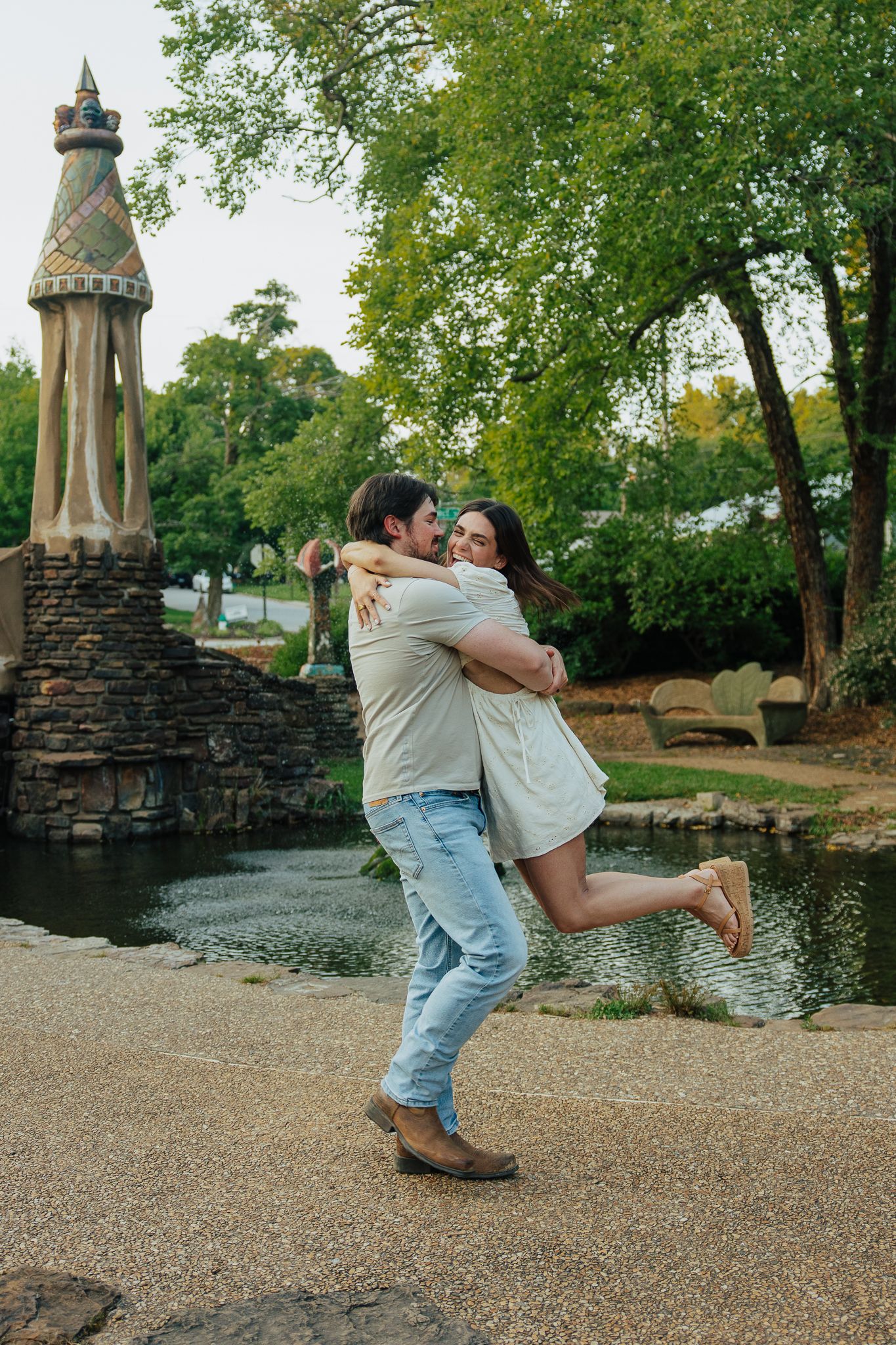 Man joyfully lifting woman in his arms beside ornate stone tower sculpture near garden pond.