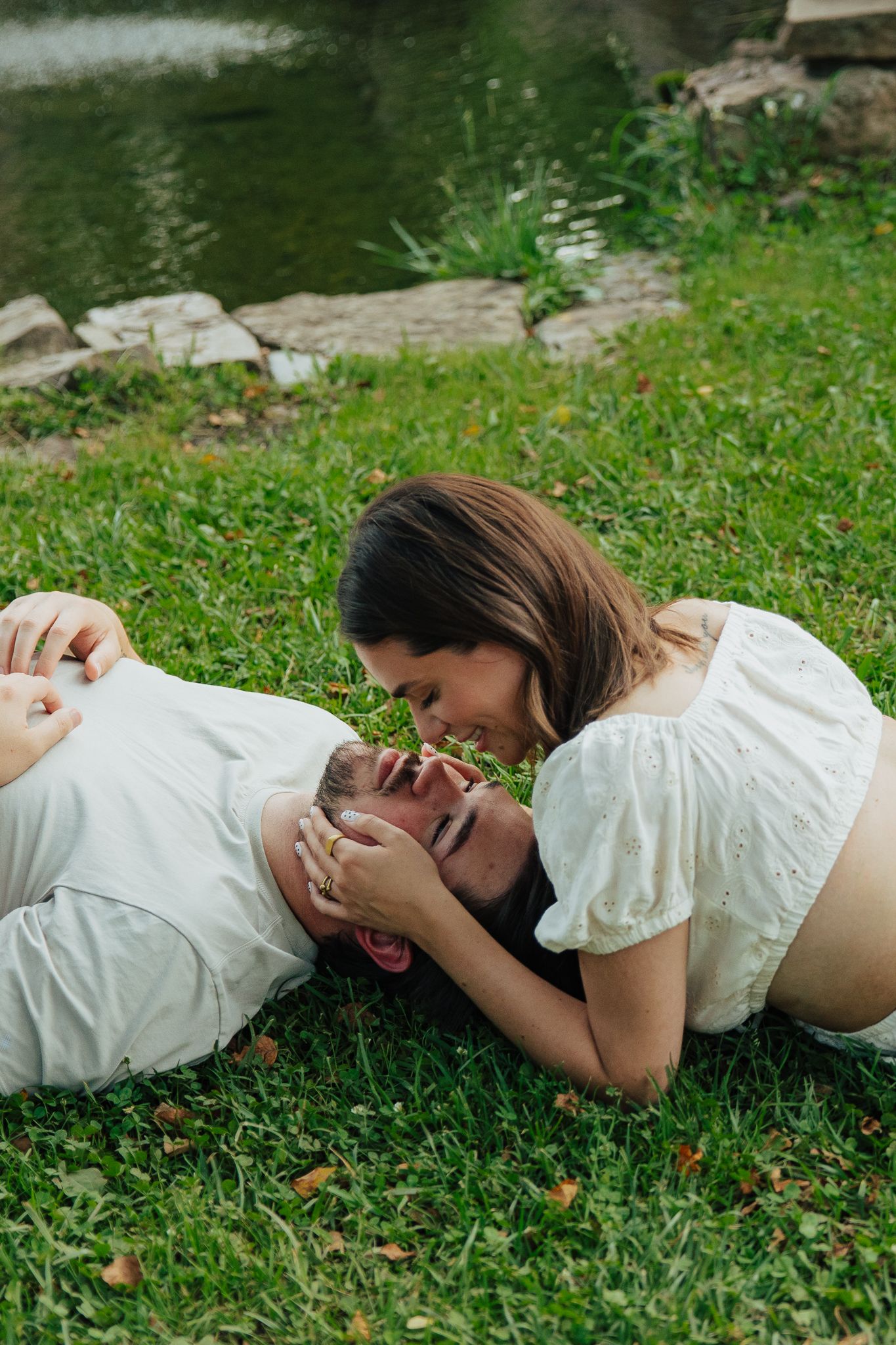 Couple lying on green grass near a pond, sharing a playful and romantic moment together.