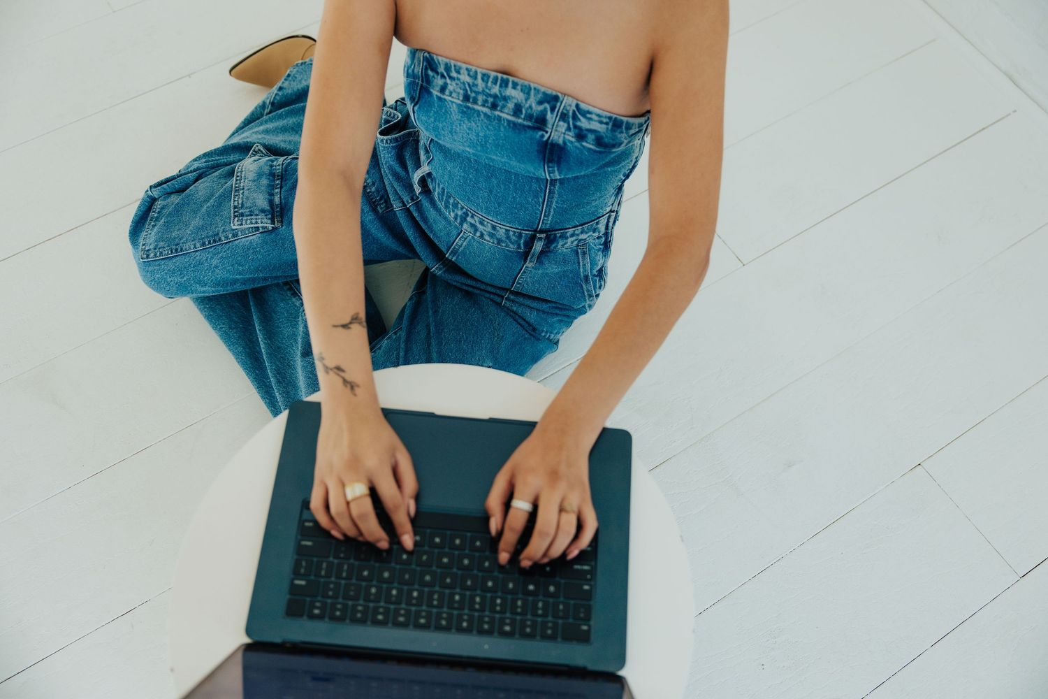 Woman in jeans with tattoo typing on laptop while sitting on white floor.